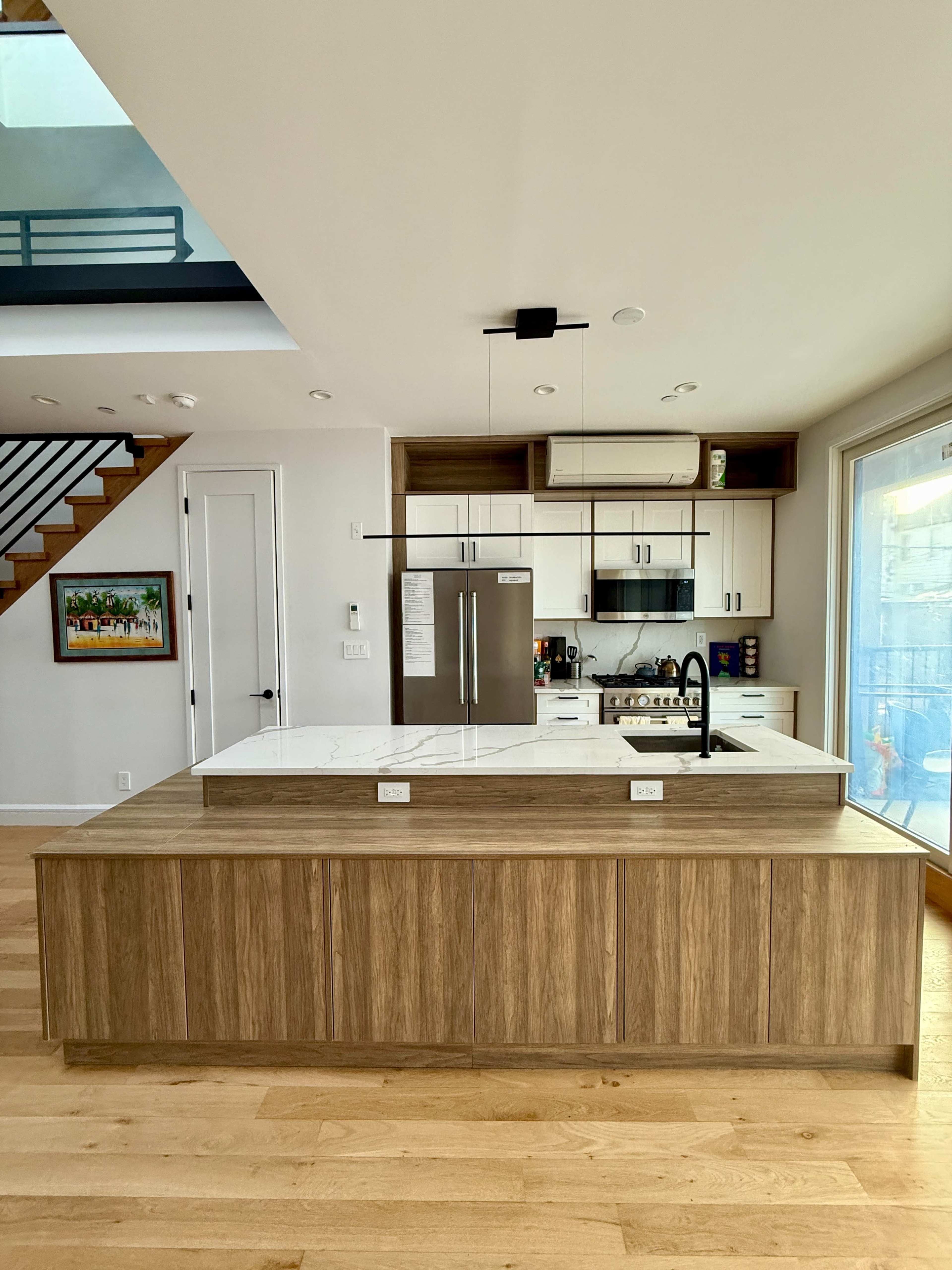 The image shows a modern kitchen with a central island made of wood, featuring a marble countertop, stainless steel appliances, and large windows providing natural light.