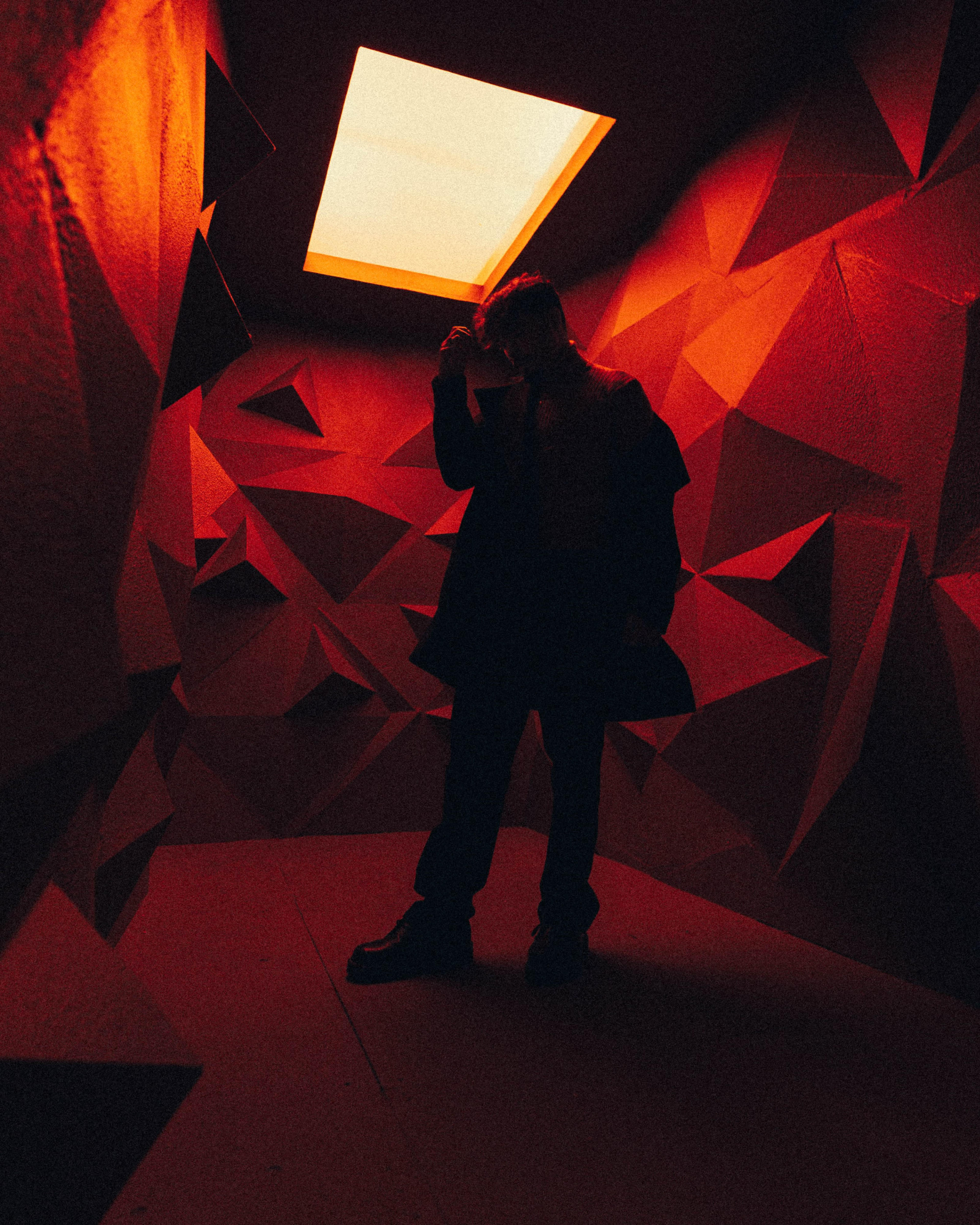 A person stands in a dimly lit, geometric room with red walls and a skylight above.