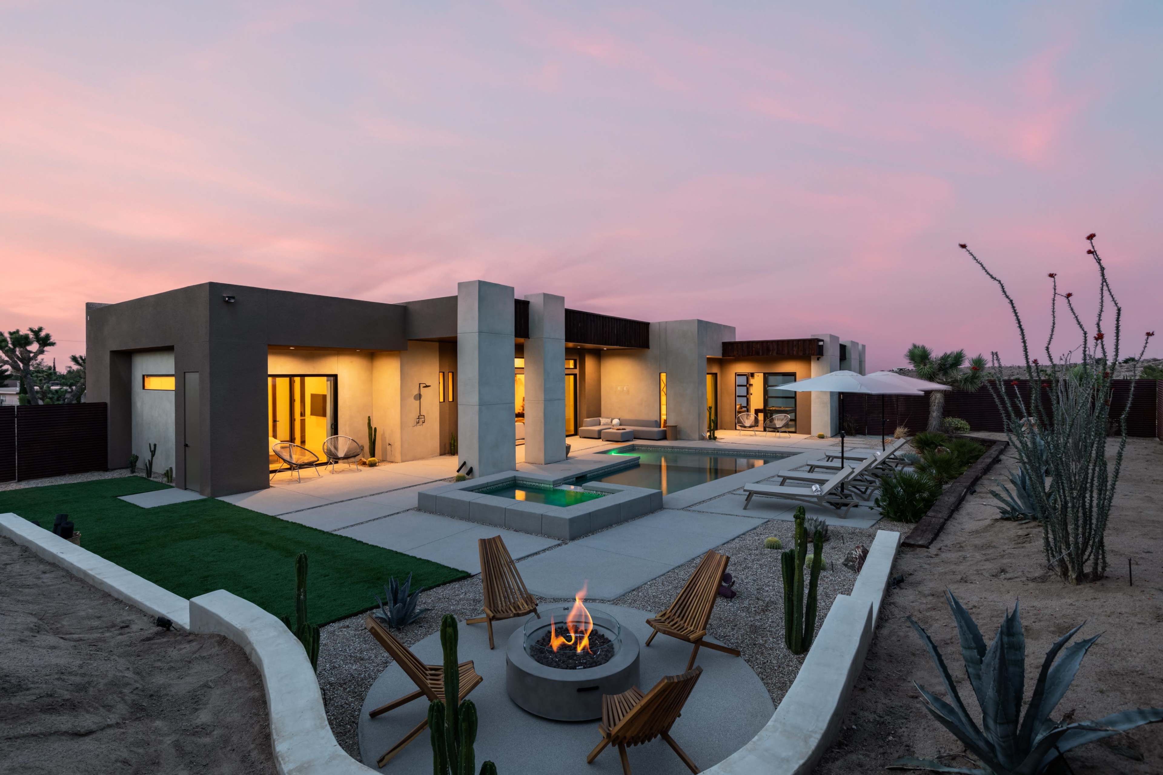 A modern desert house features a pool and outdoor seating area, with a fire pit surrounded by lounge chairs against a backdrop of a colorful sunset sky.