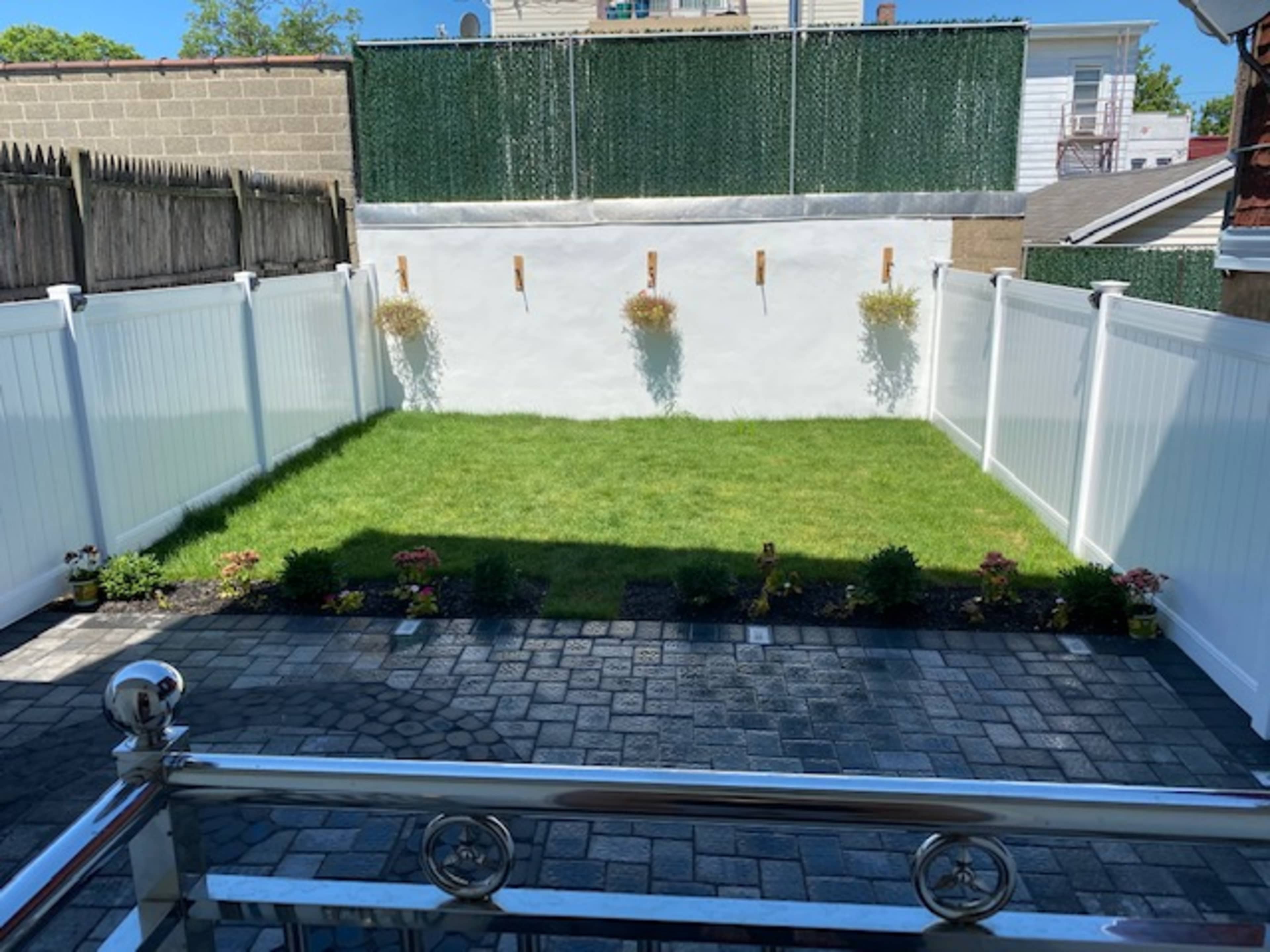 A fenced backyard with neatly trimmed grass and small flower beds along the edges, featuring white fencing and a clean stone patio.