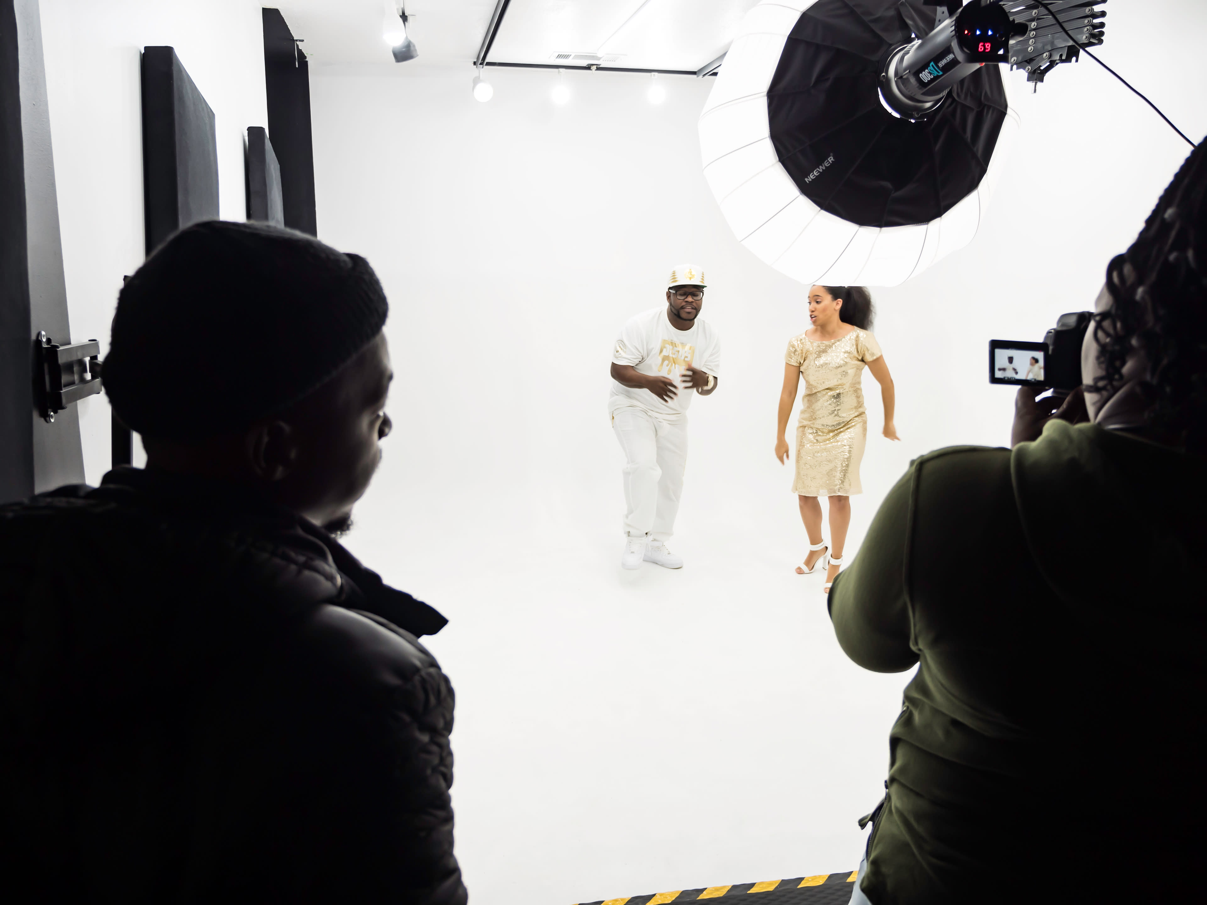 A man and a woman are posing in a photography studio with white backdrop lights while two people are observing and capturing the moment.