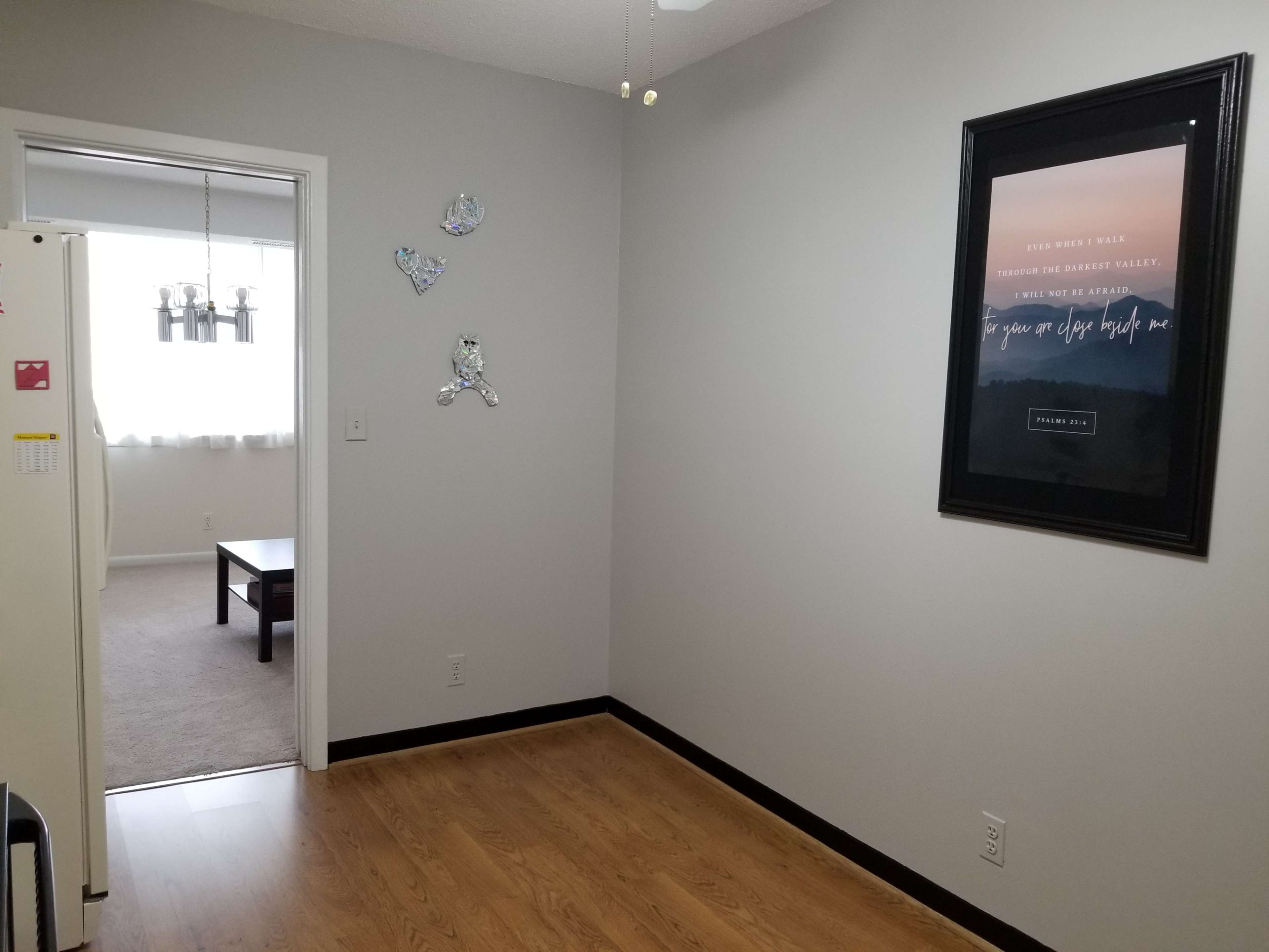A clean kitchen area features a refrigerator, an adjoining living space visible through an open doorway, and a framed motivational poster on the wall.