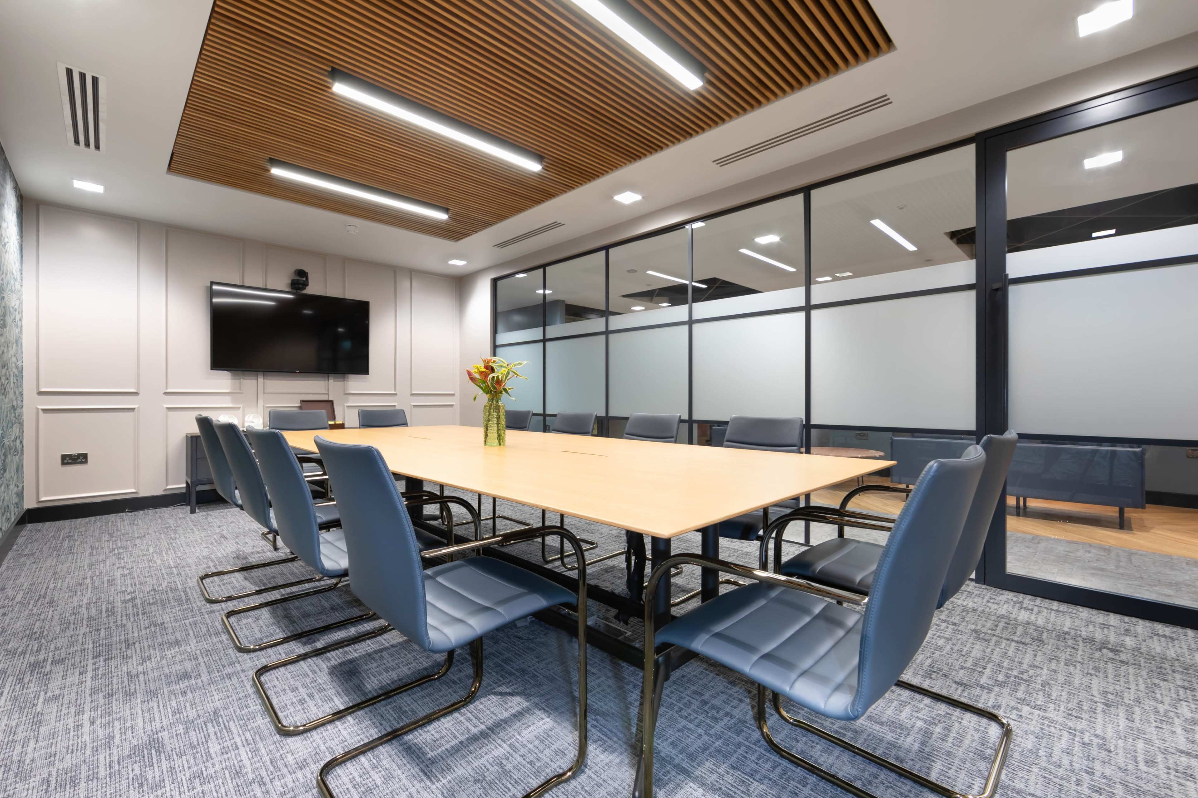 A modern conference room features a large rectangular table surrounded by metal-framed chairs, with glass walls and a television mounted on one side.