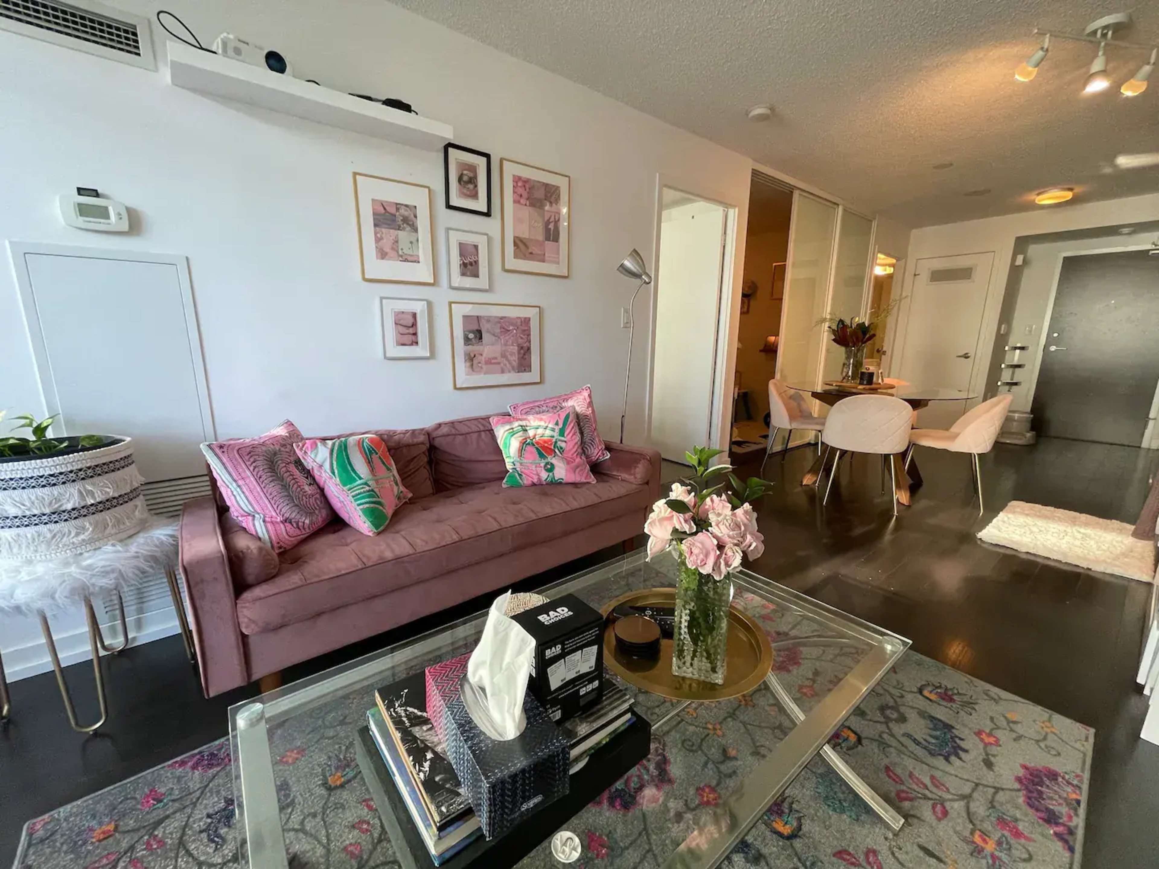 A modern living room features a pink sofa, decorative art on the walls, a coffee table with flowers, and a dining area visible in the background.