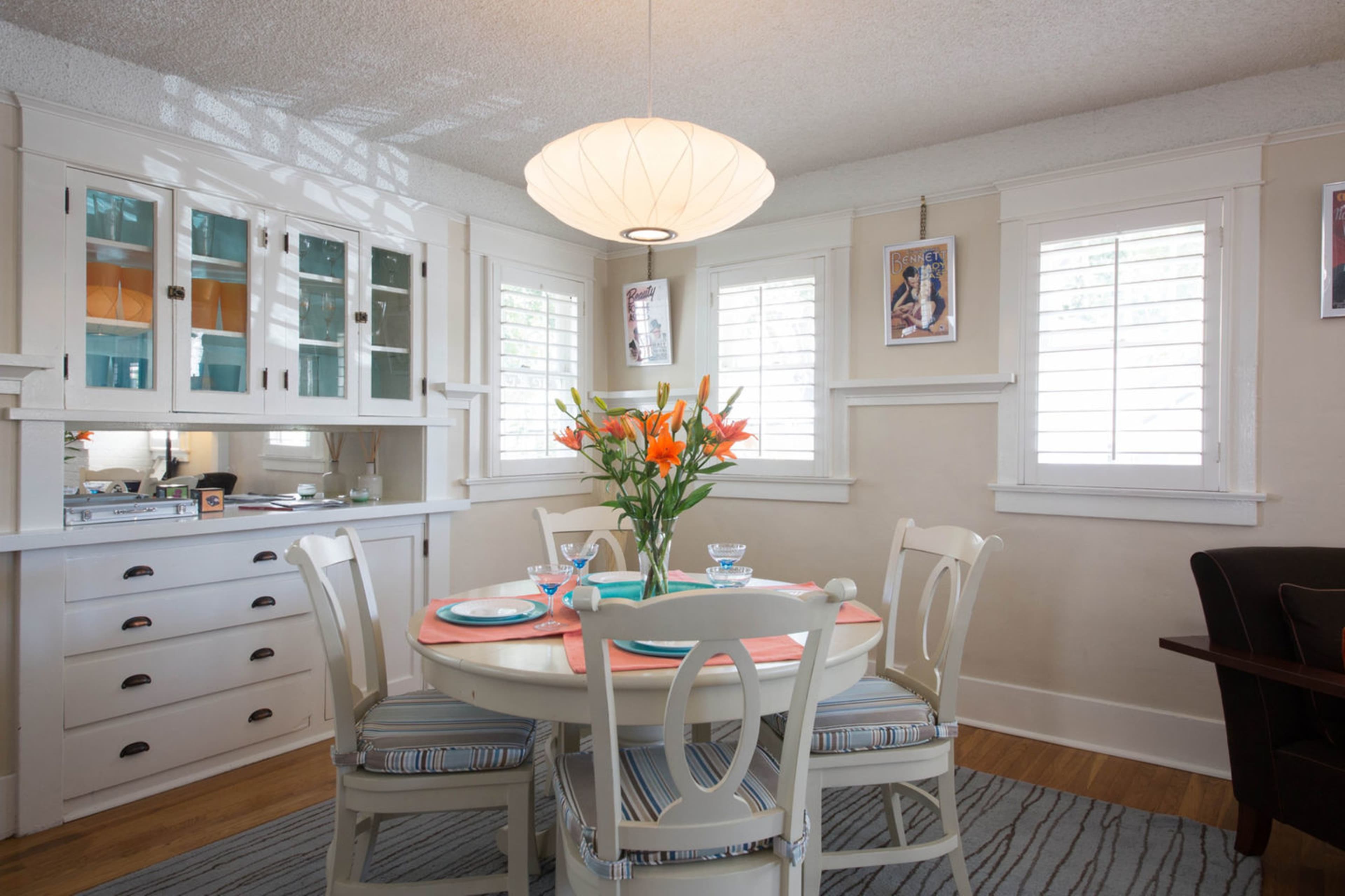 The image shows a dining area with a round table set for four, surrounded by white chairs and adorned with a vase of orange flowers, with a light fixture hanging above.