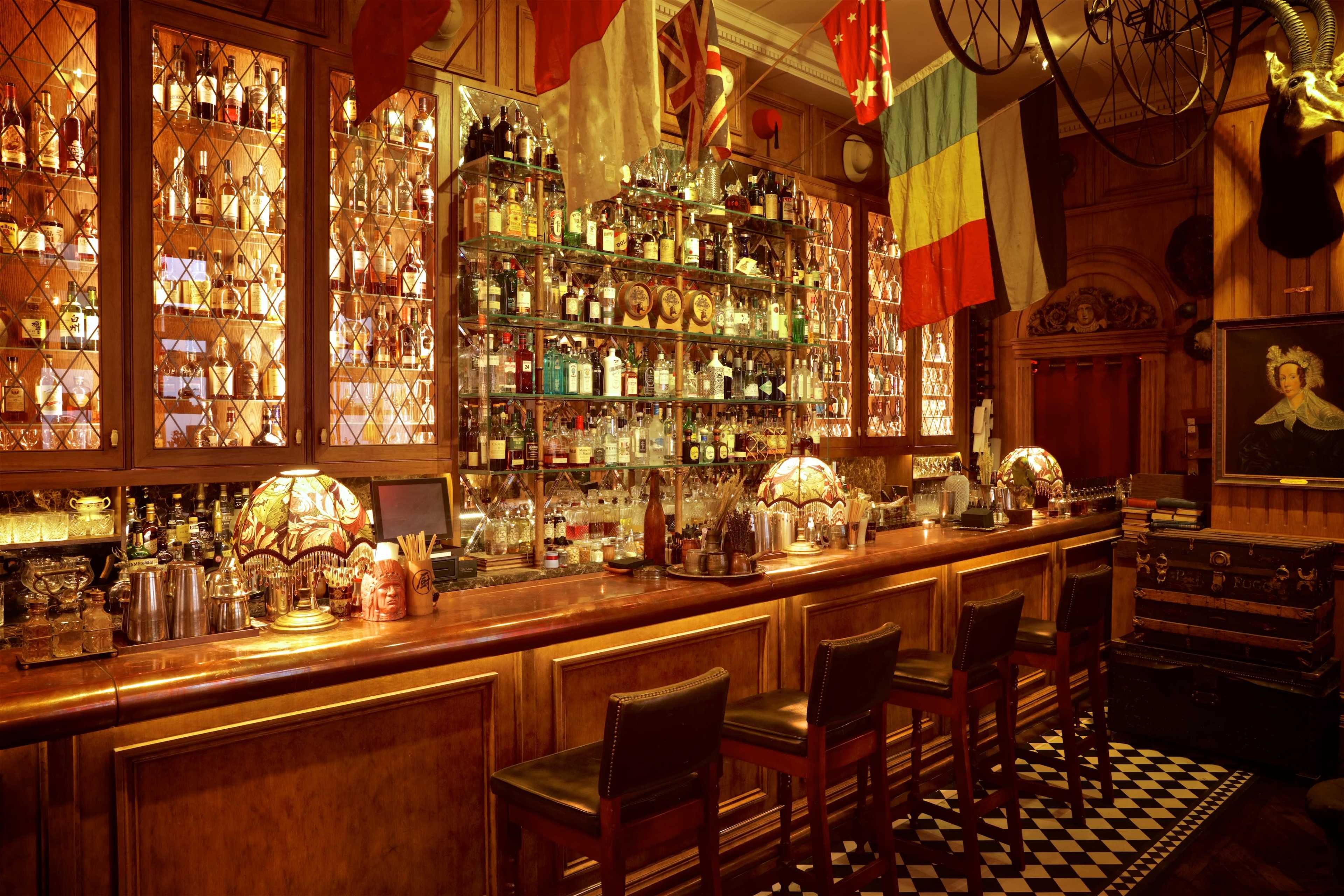 The image shows a well-stocked wooden bar with a mirrored backlit shelf displaying various bottles, surrounded by flags and illuminated by decorative lamps.