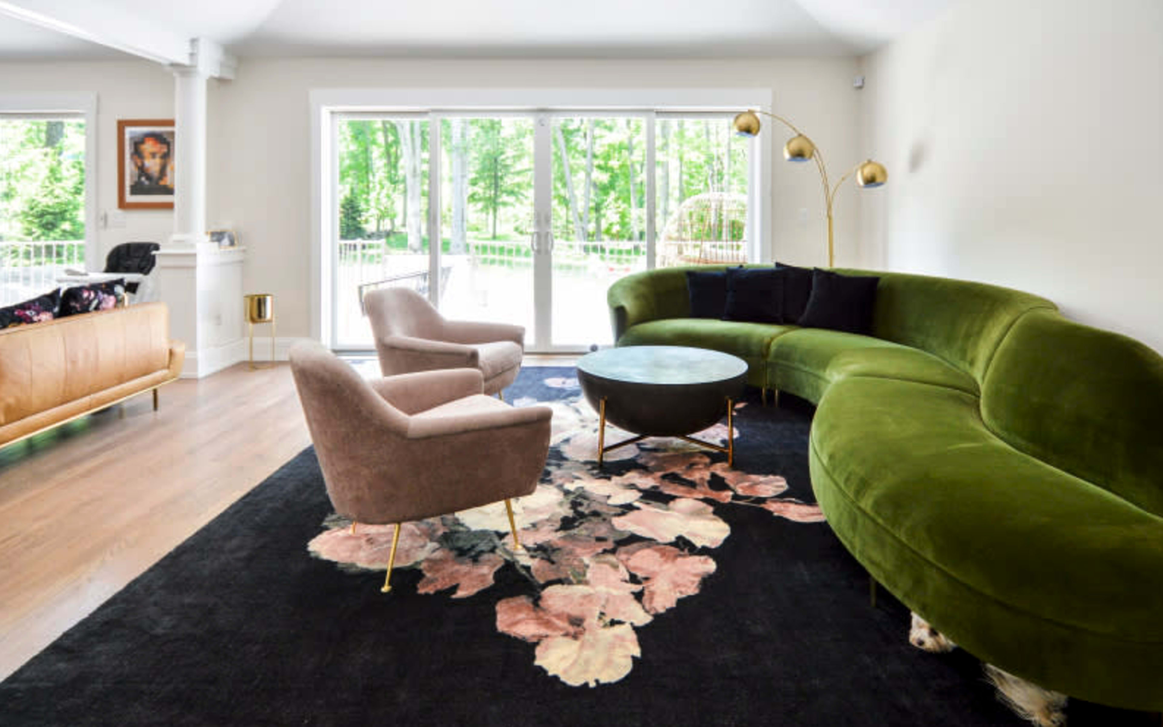 The image shows a modern living room featuring a green curved sofa, two light-colored armchairs, a round coffee table, and a dark floral-patterned rug, with large windows overlooking a green outdoor area.