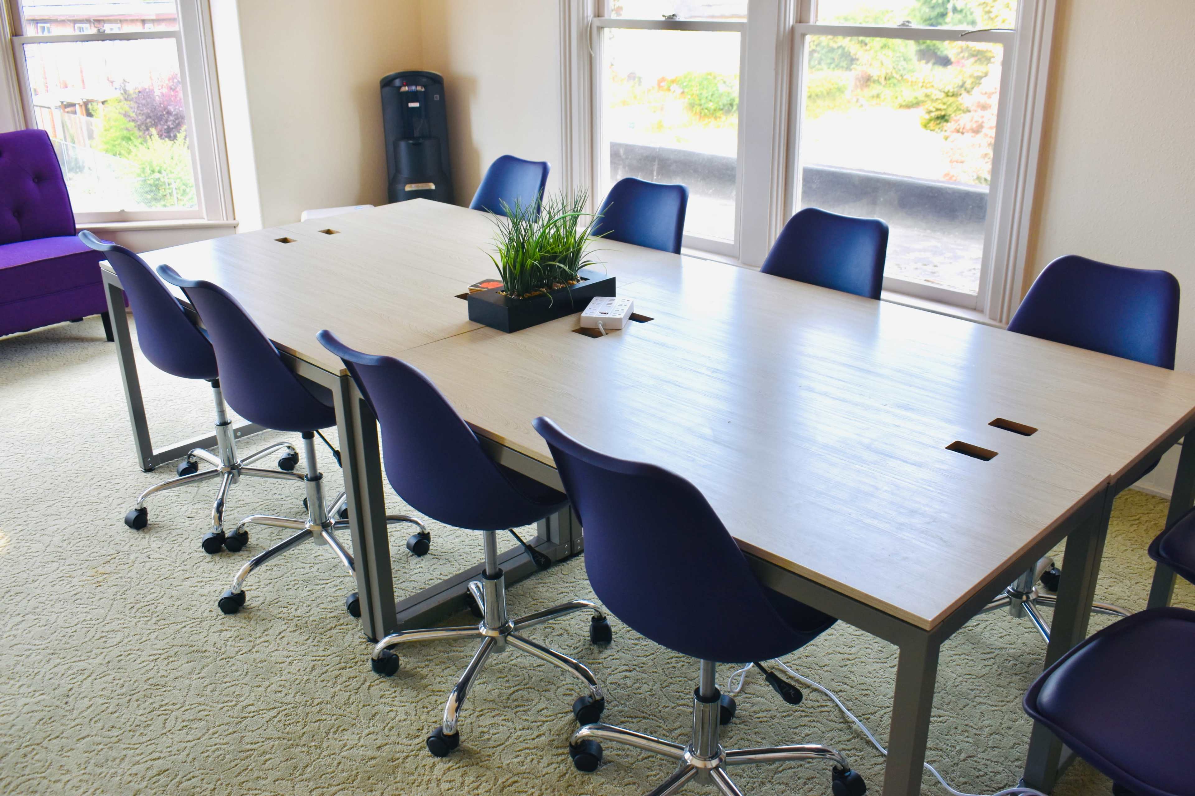 A large meeting table surrounded by purple rolling chairs is set in a well-lit room with windows overlooking greenery.