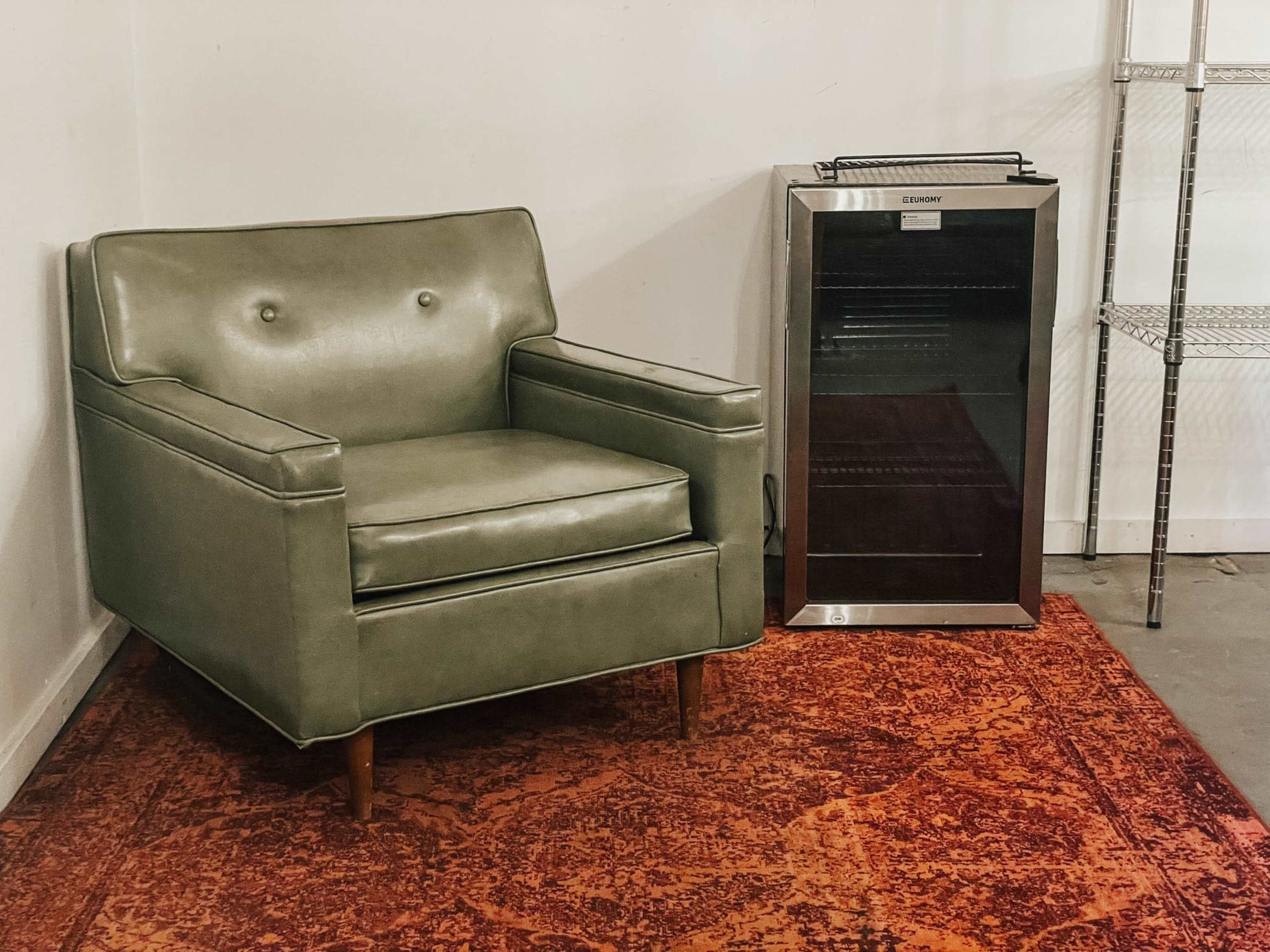 A green upholstered armchair sits next to a black beverage fridge on a patterned red rug in a minimalist room.