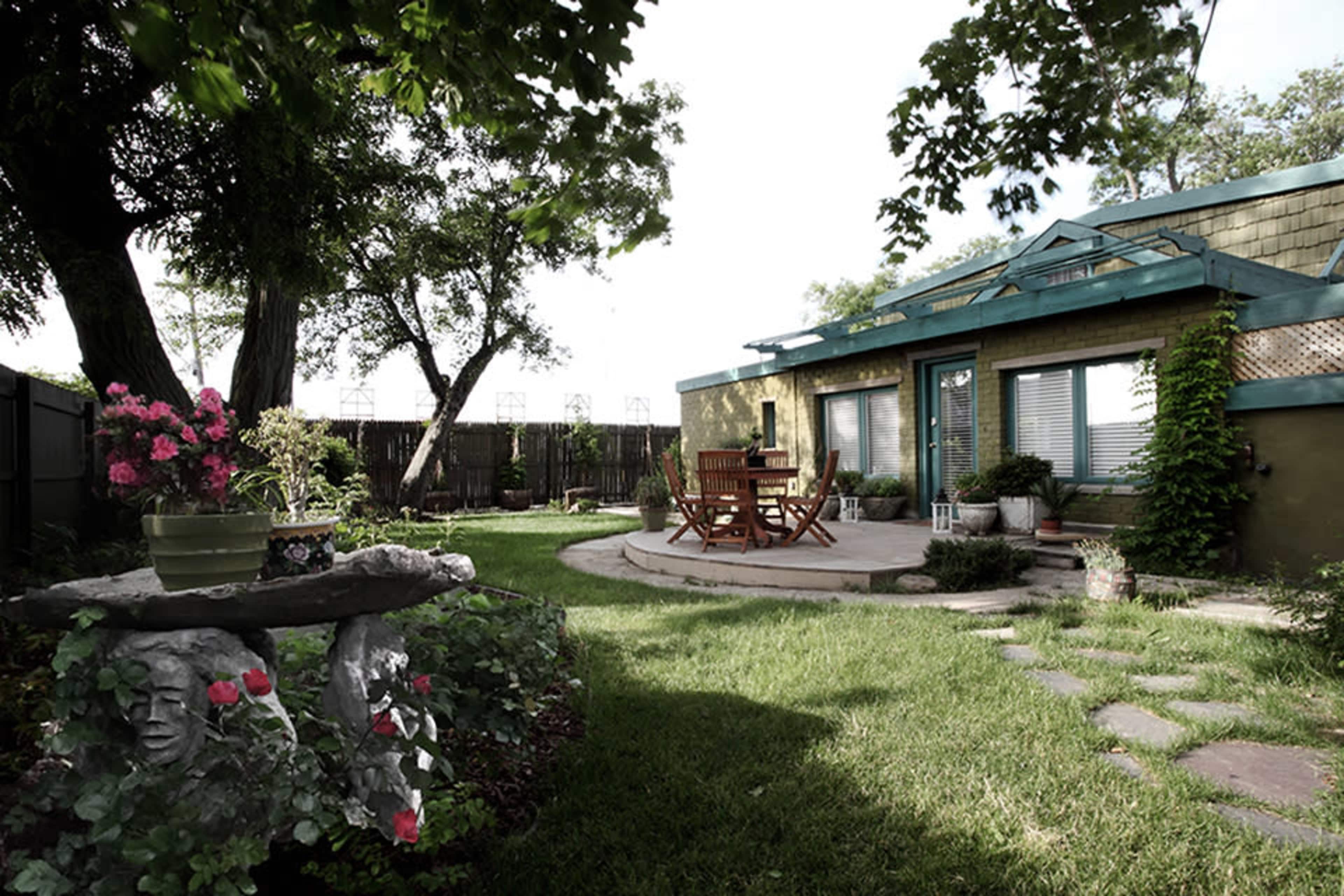 A landscaped backyard with a stone table, flower pots, a wooden dining set, and a small patio area adjacent to a house.