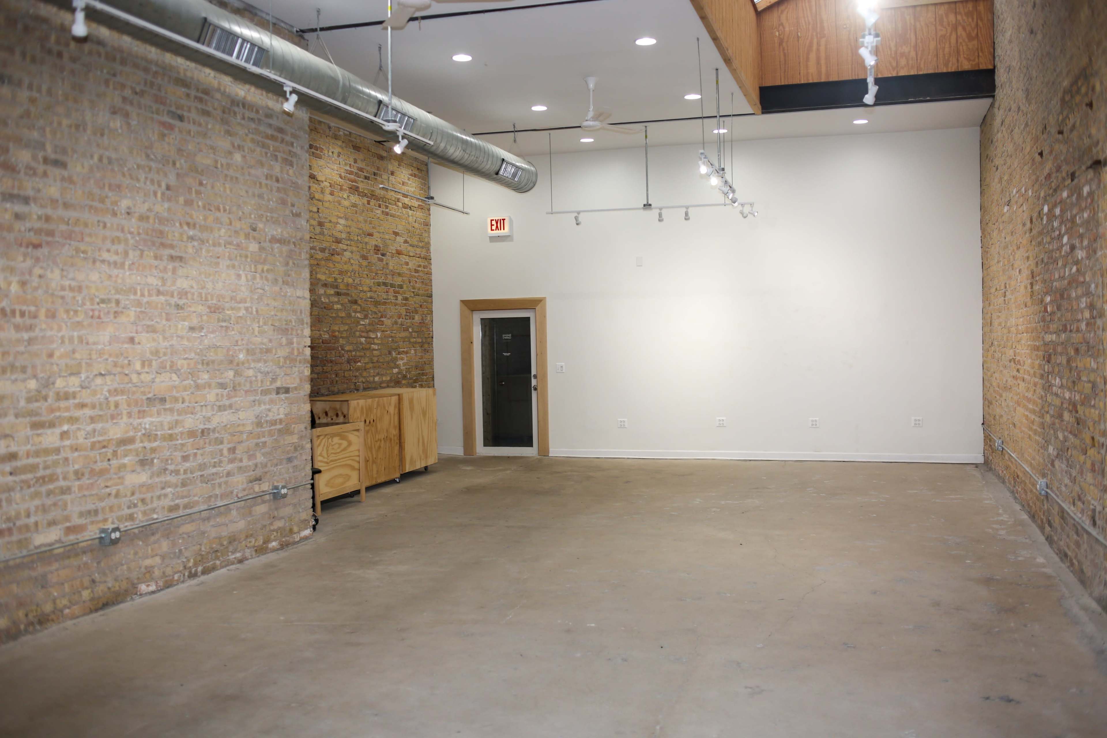 The image shows a vacant room with exposed brick walls, a concrete floor, and overhead lighting, featuring an entrance door on one side and a small kitchenette against one wall.