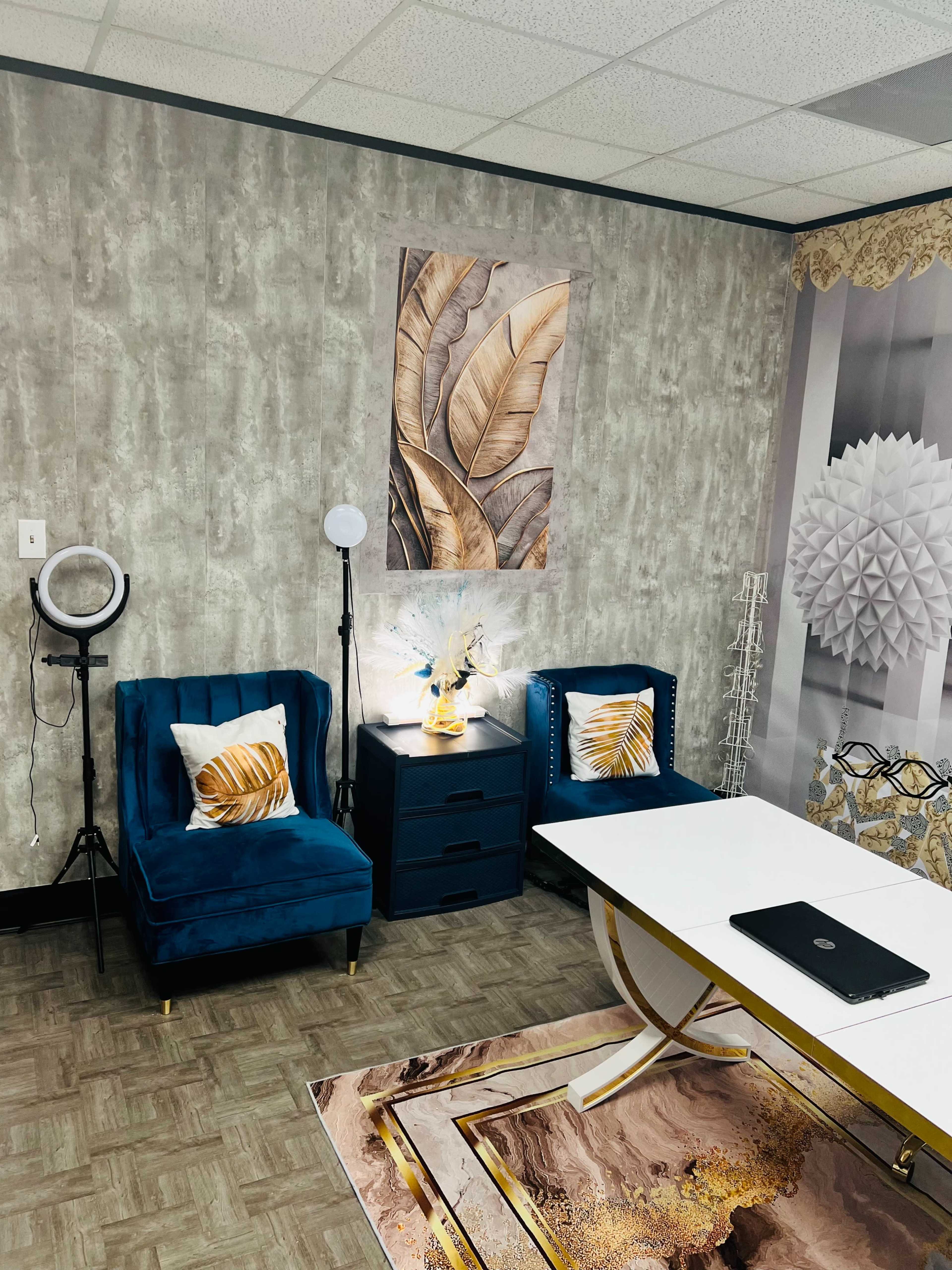 The image shows a modern office space featuring two blue velvet chairs, a side table with a decorative lamp and plant, and a large artwork depicting leaves on the wall.