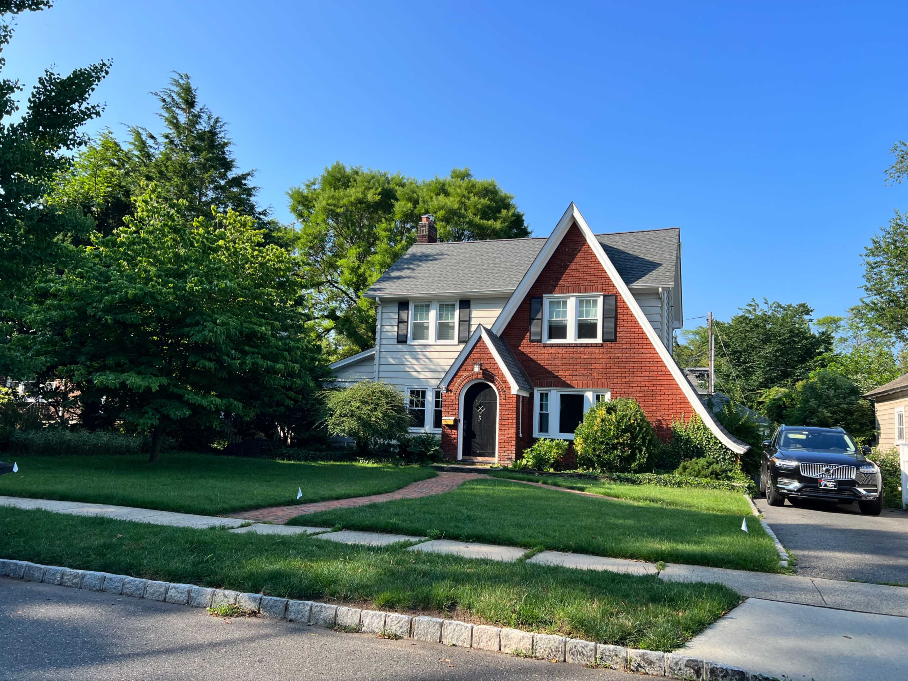A two-story, red-brick house with white trim and a steeply pitched roof, set in a landscaped yard with green grass and trees.