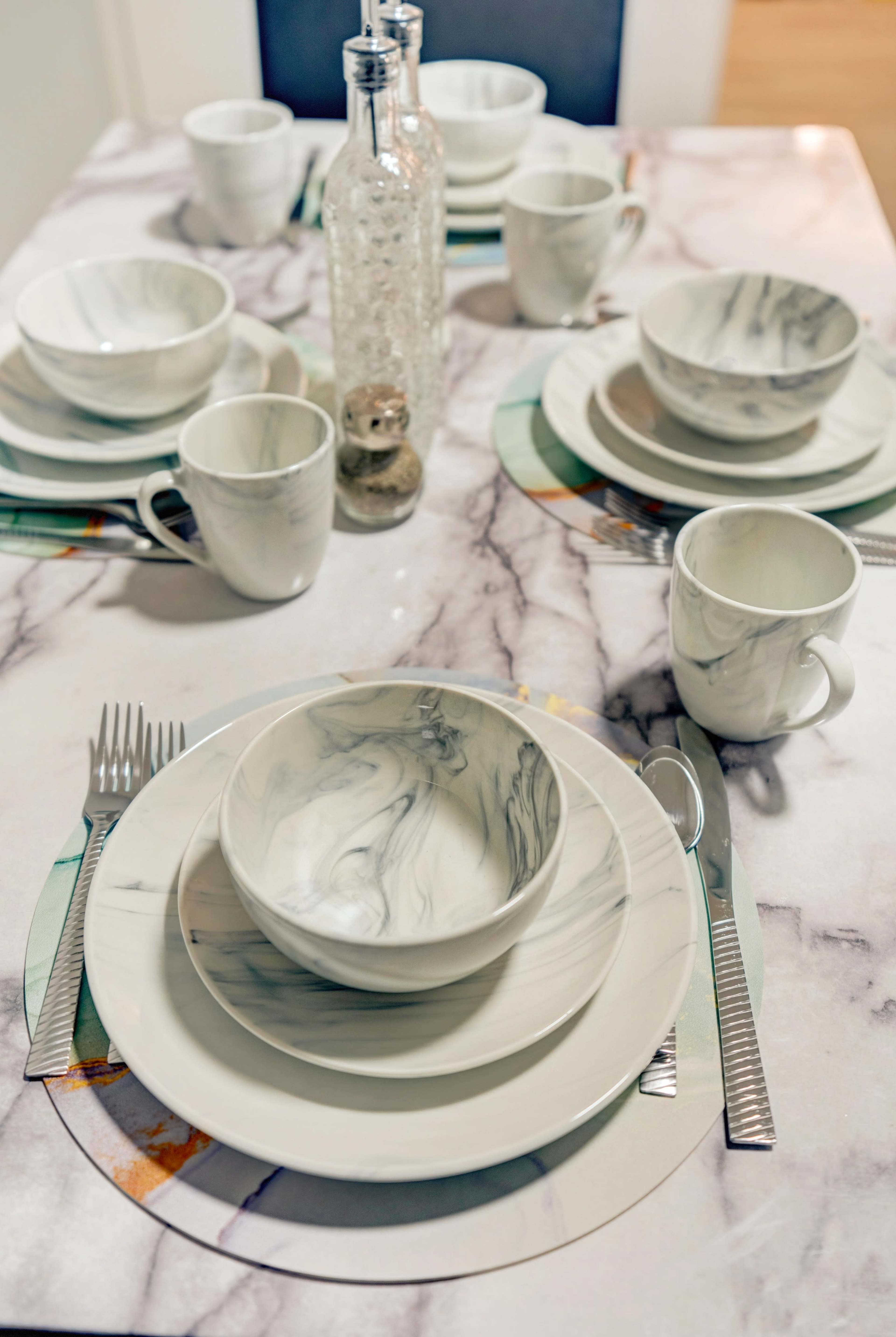 A dining table is set with marble-patterned dishware, including bowls, plates, and cups, arranged neatly with silver utensils and a decorative glass bottle centerpiece.