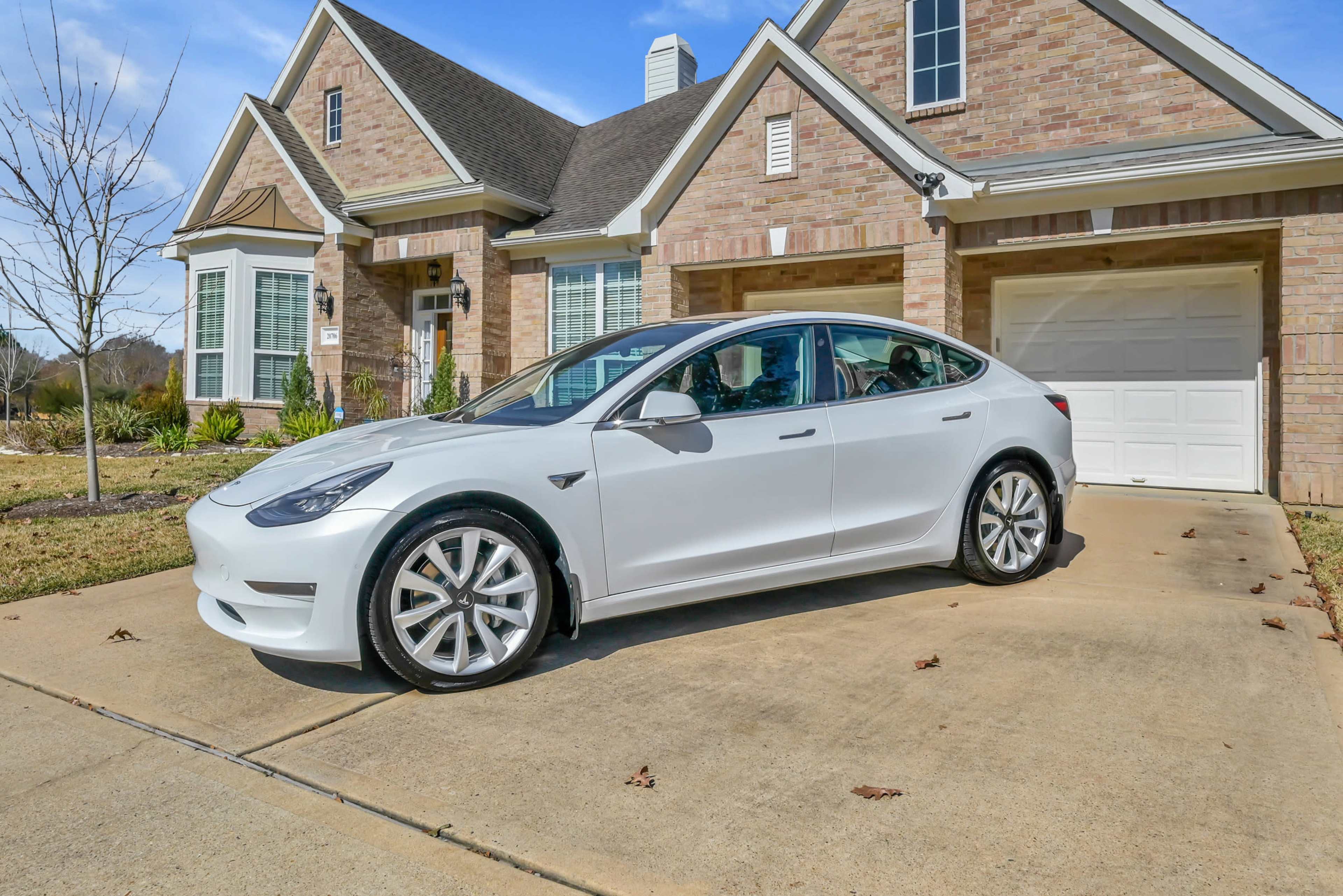 A white Tesla Model 3 is parked in the driveway of a brick house on a sunny day.