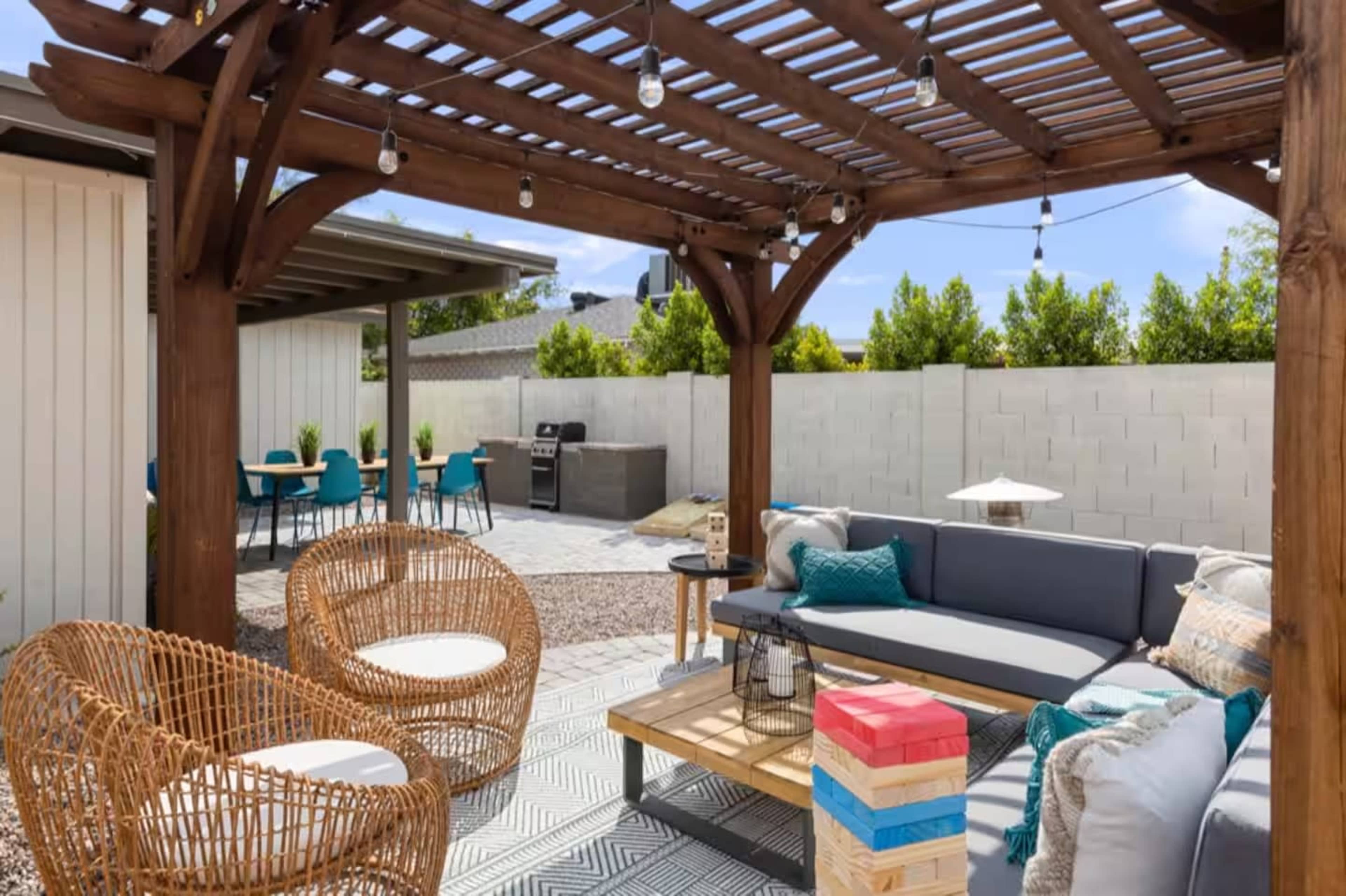 The image shows a shaded outdoor patio area featuring wooden furniture, a seating arrangement, and a barbecue grill in the background.