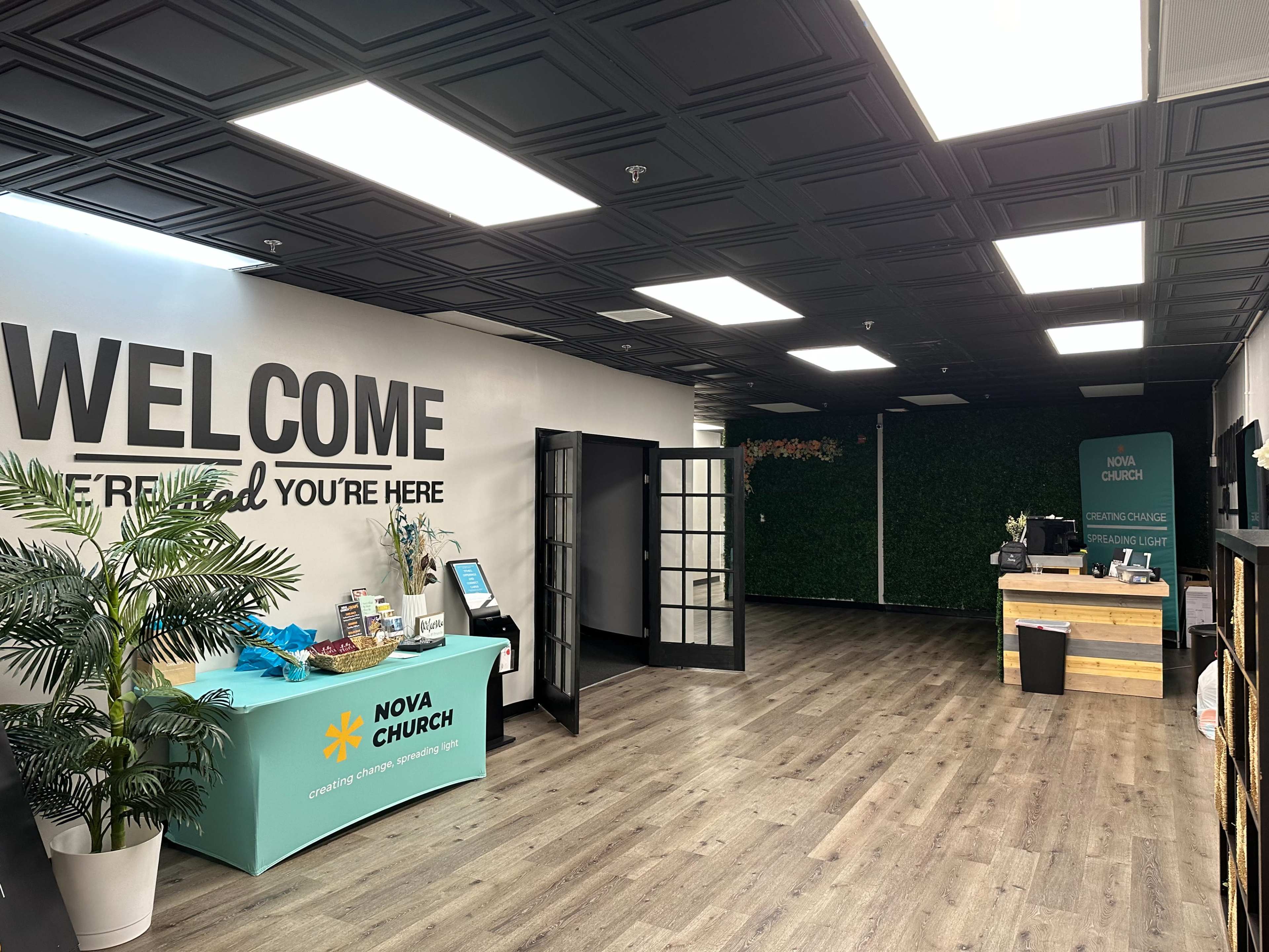 The interior of a church lobby features a welcome sign, a reception desk, and a green wall with plants.