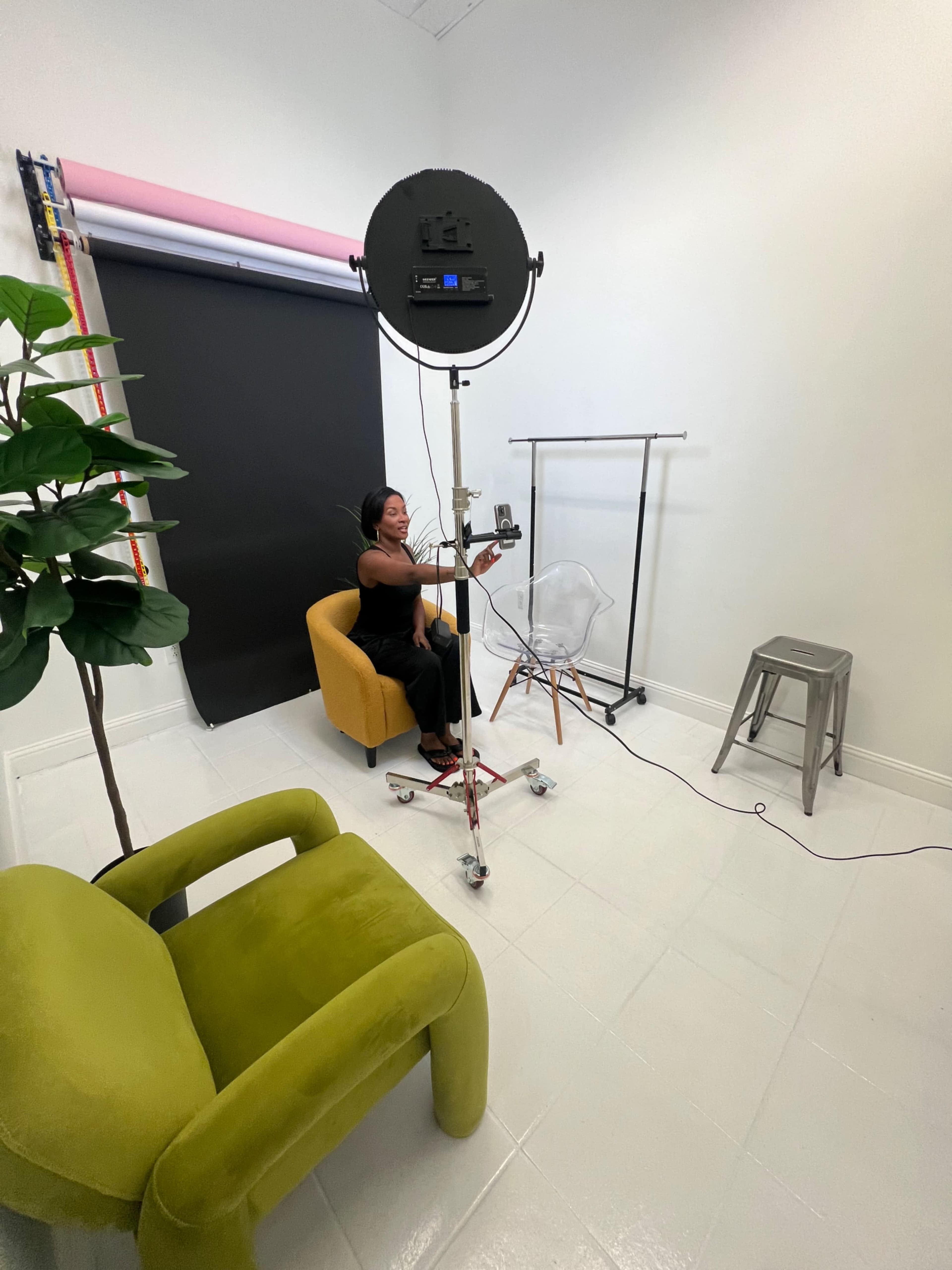 A woman is seated in an orange chair in a brightly lit photo studio, adjusting a camera on a tripod while a backdrop and additional seating are positioned nearby.