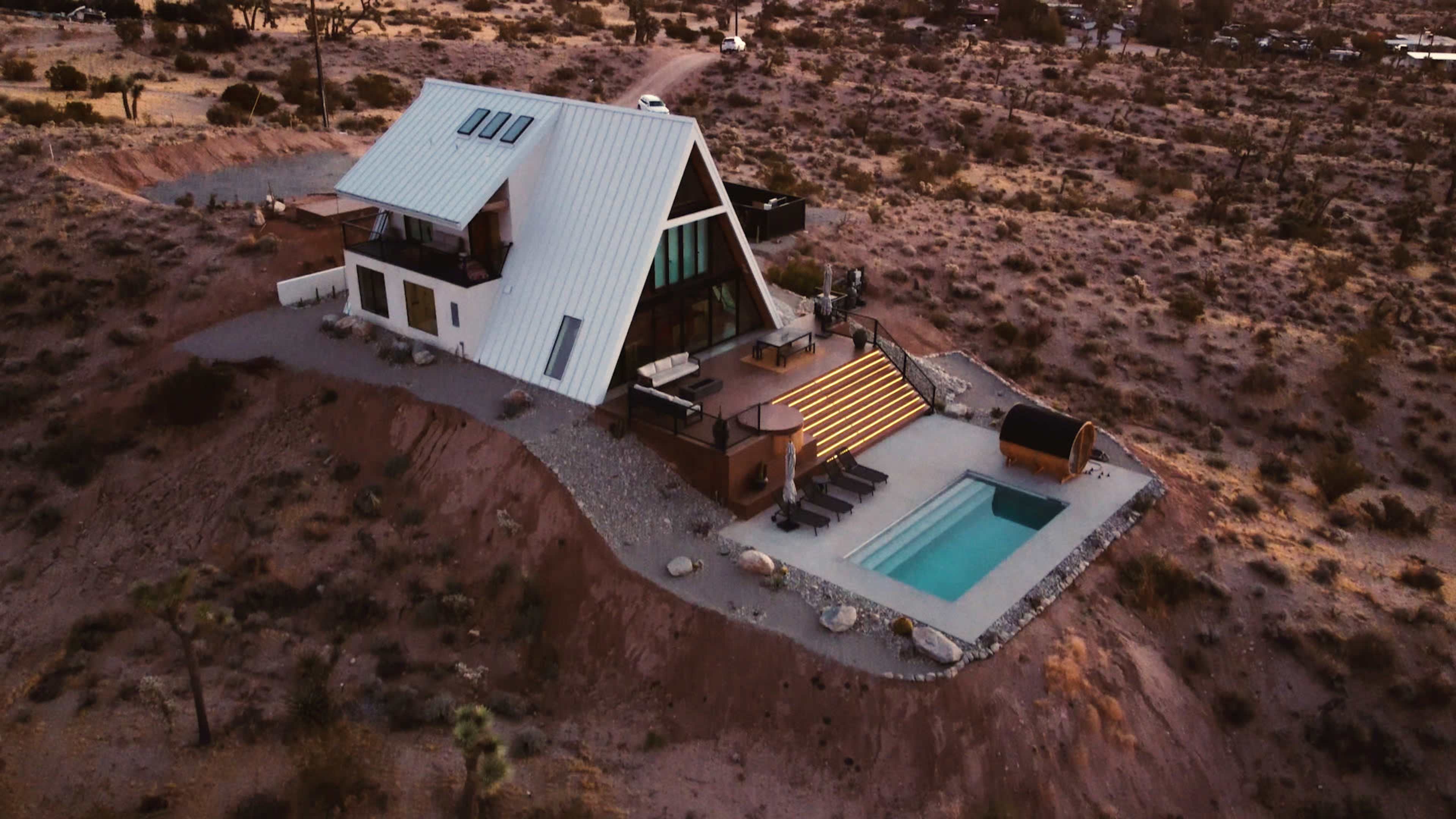 A modern A-frame house on a rocky hillside, featuring a pool and multiple outdoor spaces in a desert landscape.