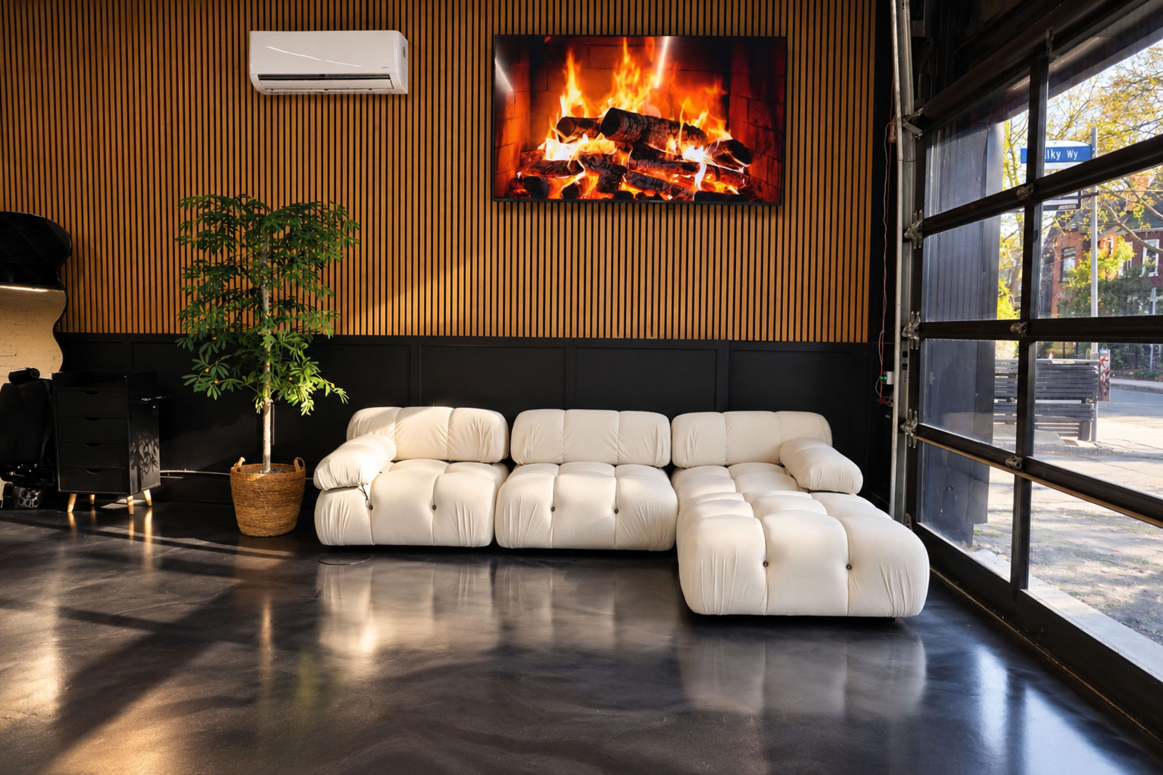A white sectional sofa is positioned against a wood-panelled wall, with a television displaying a fireplace above it, and a potted plant nearby.