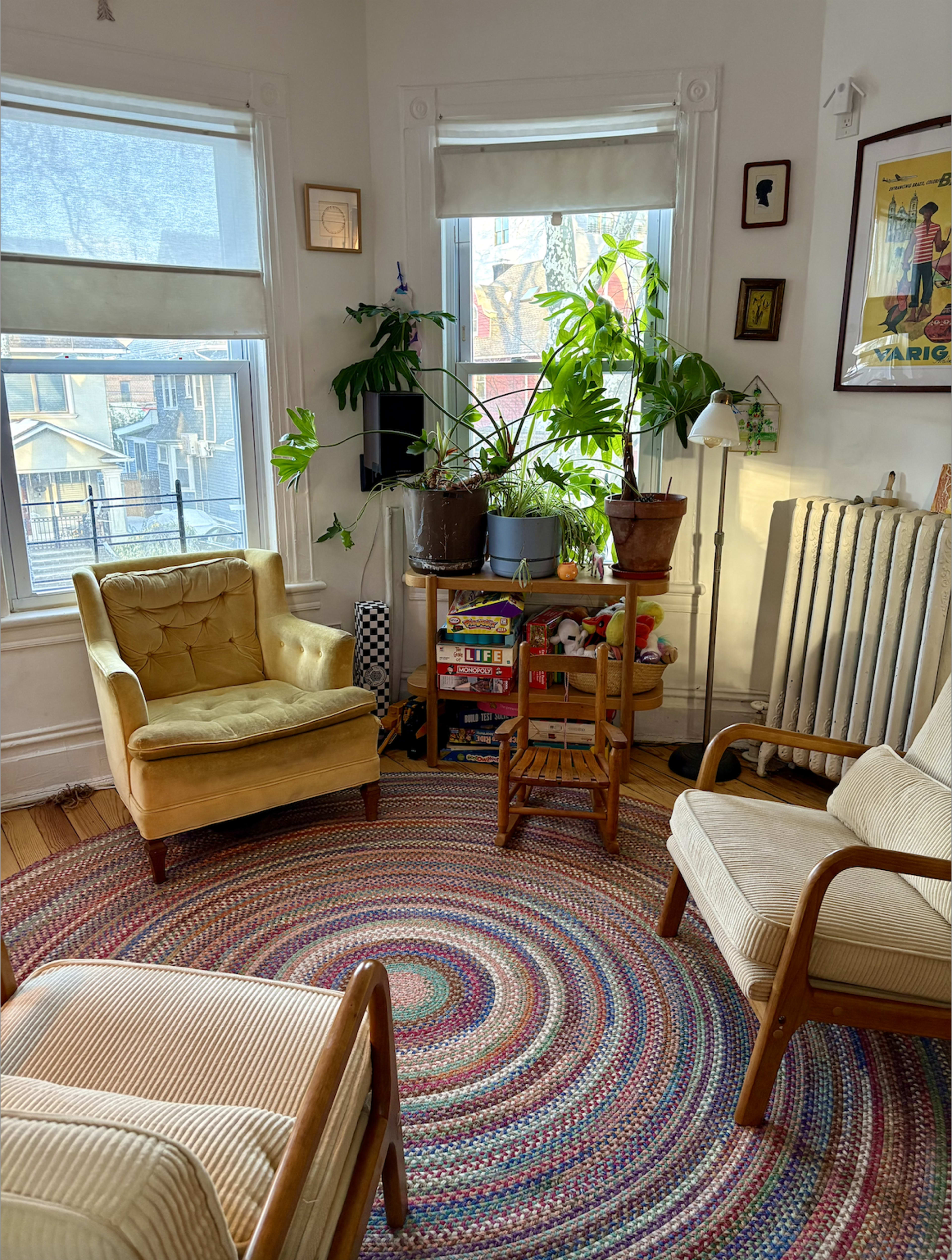 The image shows a cozy living room with two vintage armchairs, a round braided rug, and several plants near a window with sunlight streaming in.
