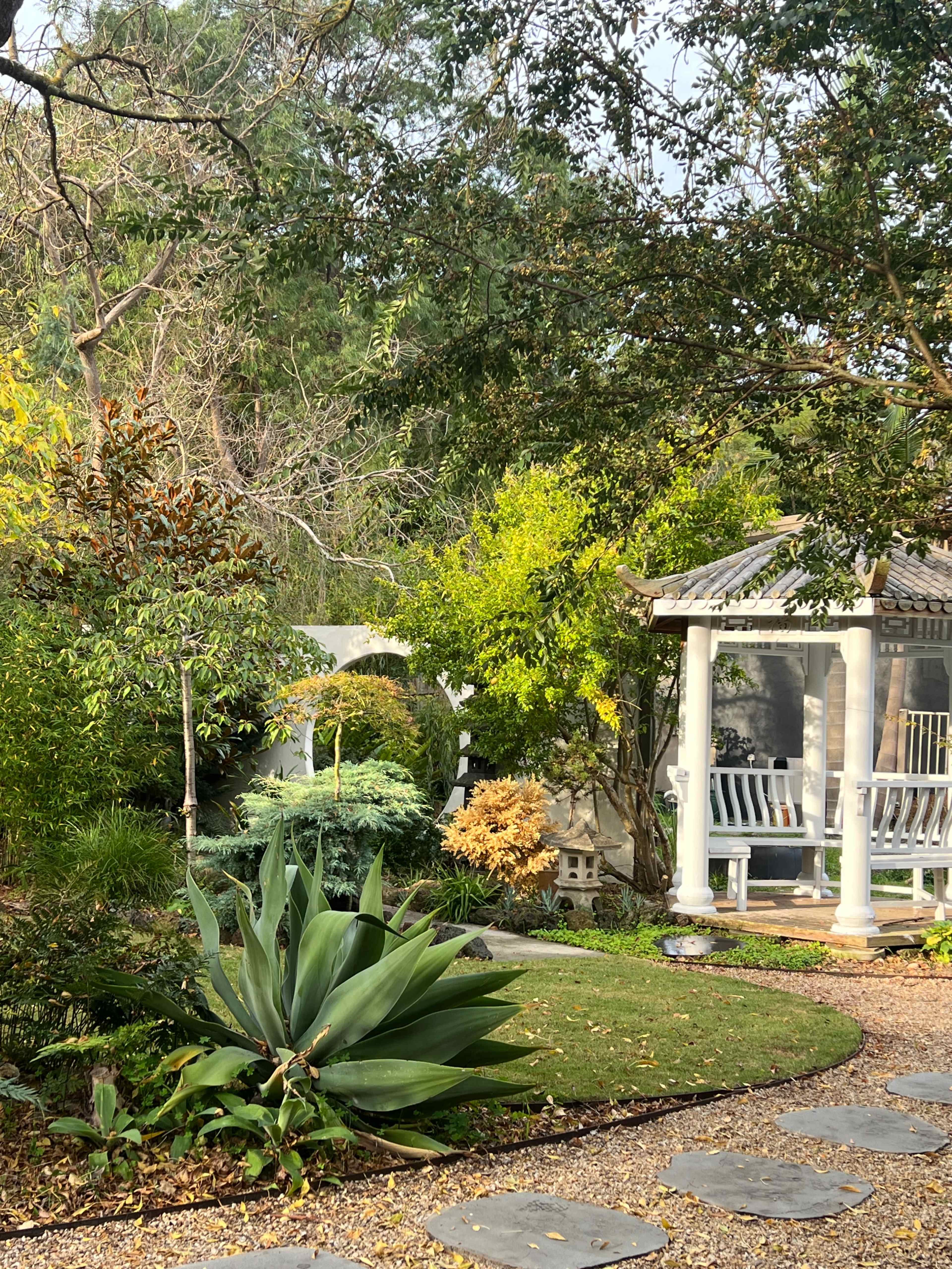 A serene garden features a stone pathway leading to a gazebo surrounded by various plants and trees.