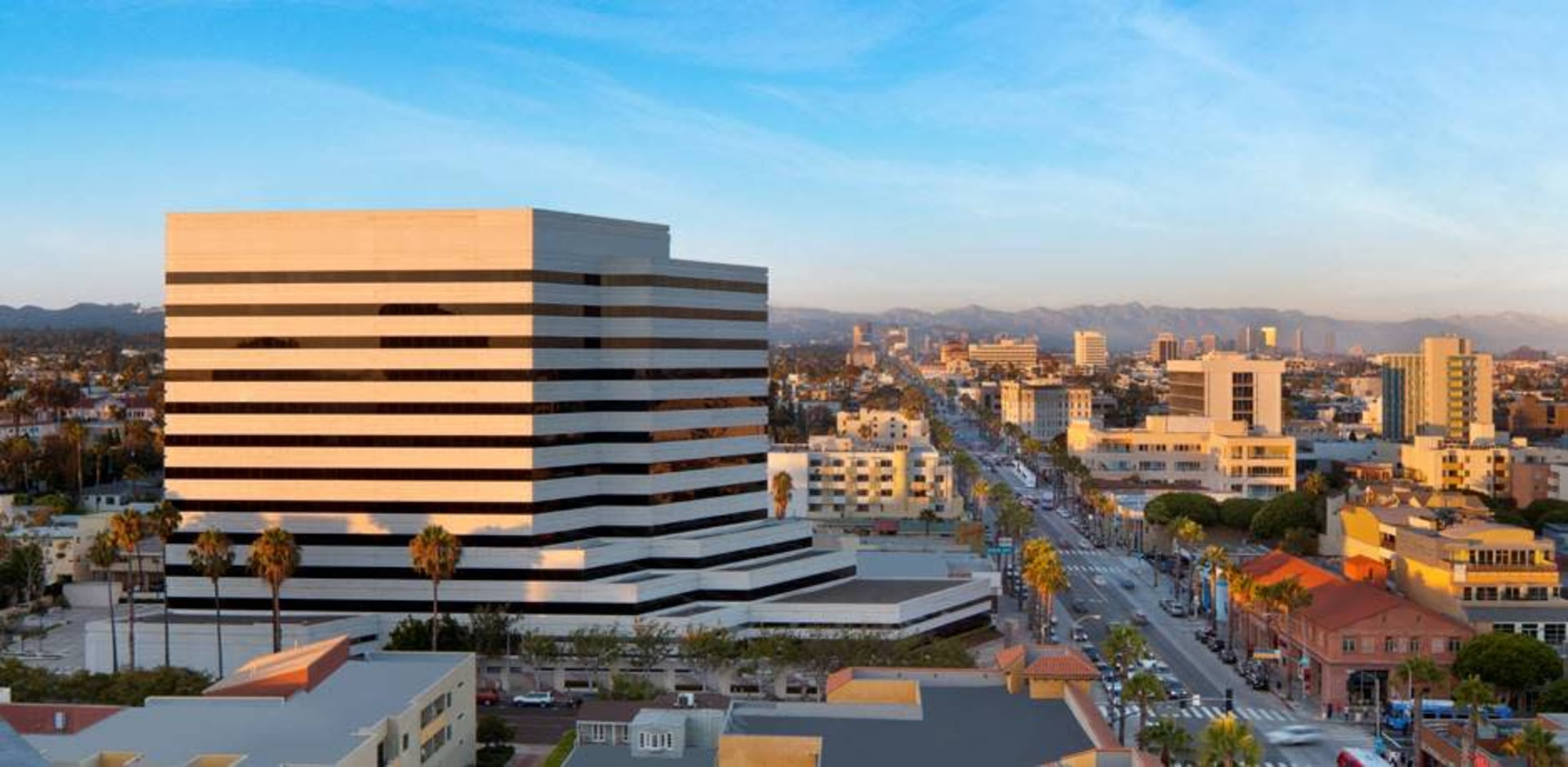 A modern, multi-story building with black and white stripes overlooks a city skyline and street filled with palm trees and various commercial buildings.