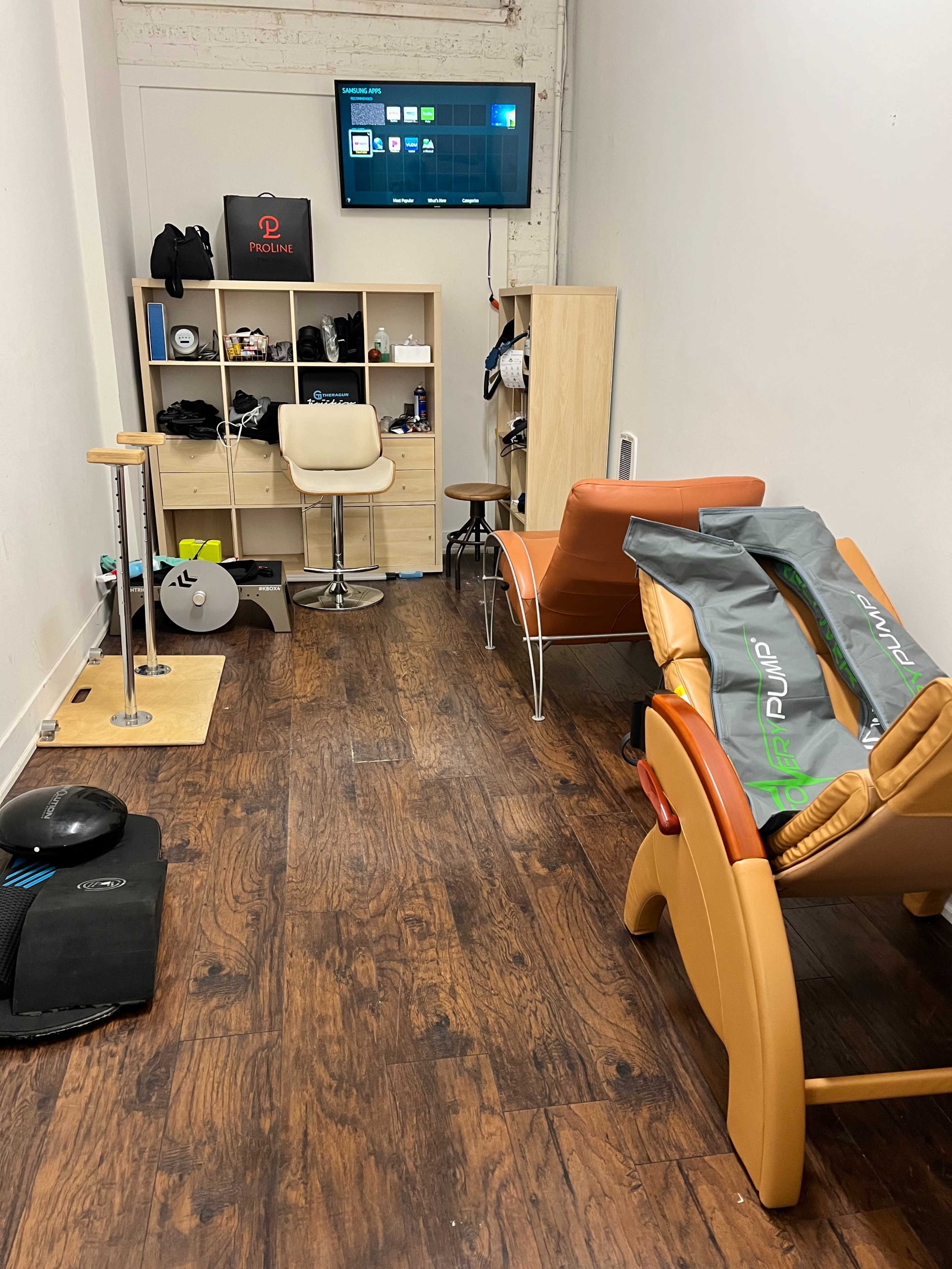 A narrow room with wooden flooring, featuring a TV mounted on the wall, shelves filled with equipment, and two lounge chairs.