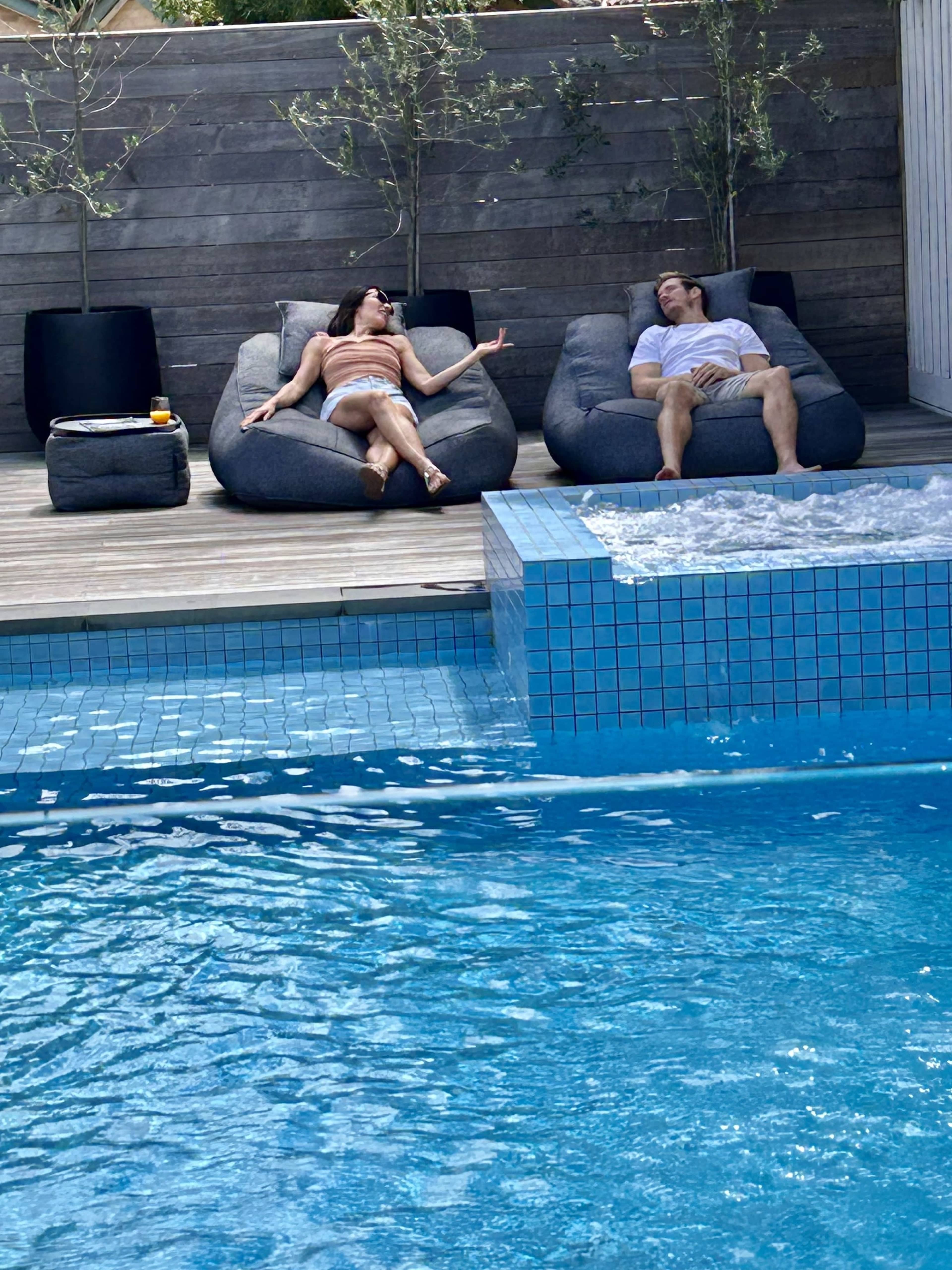 Two people relax on bean bag chairs beside a swimming pool with a small spa area.