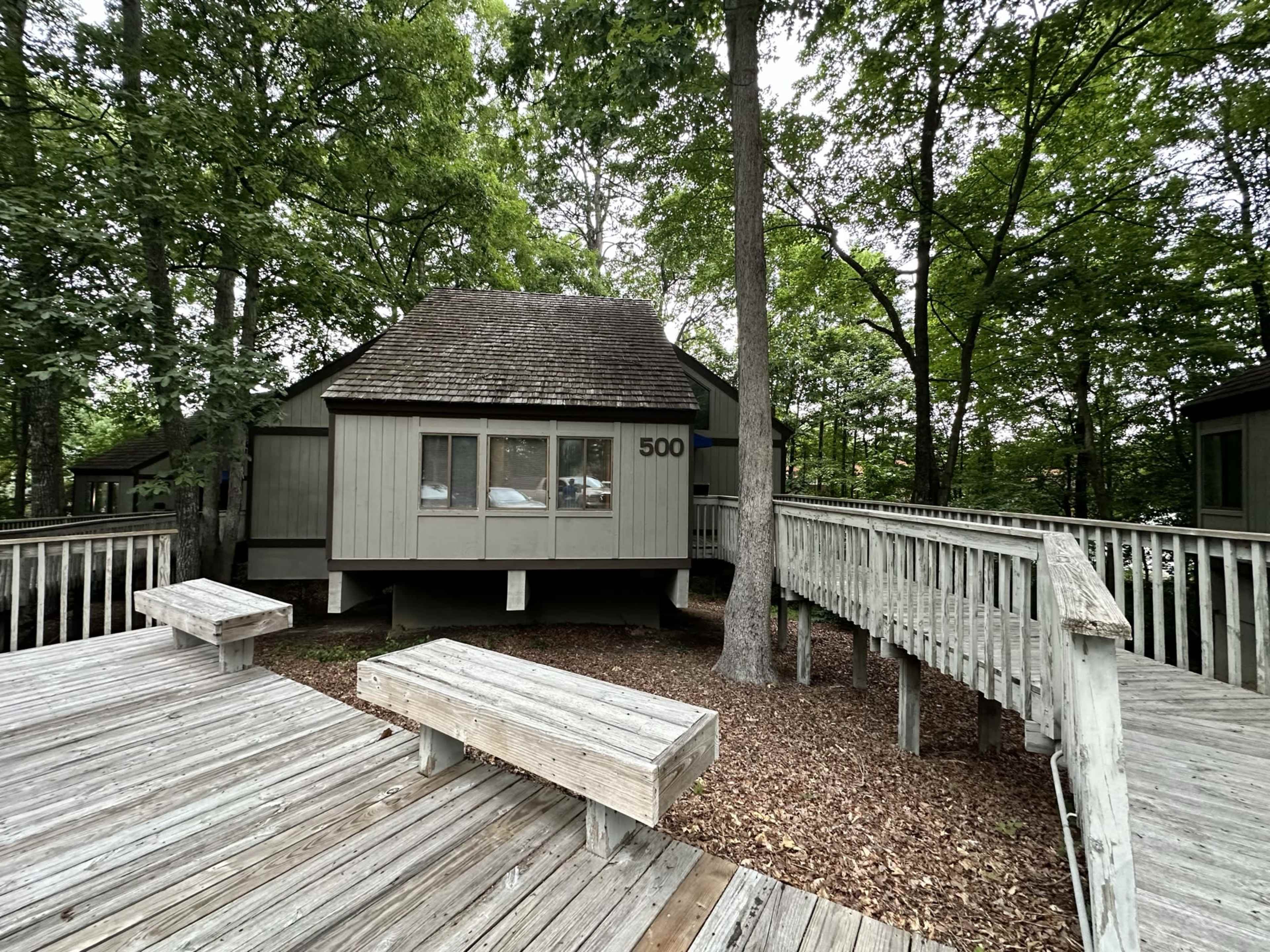 A cabin with the number 500 is situated among trees, connected by wooden walkways and surrounded by a deck area covered in wood chips.
