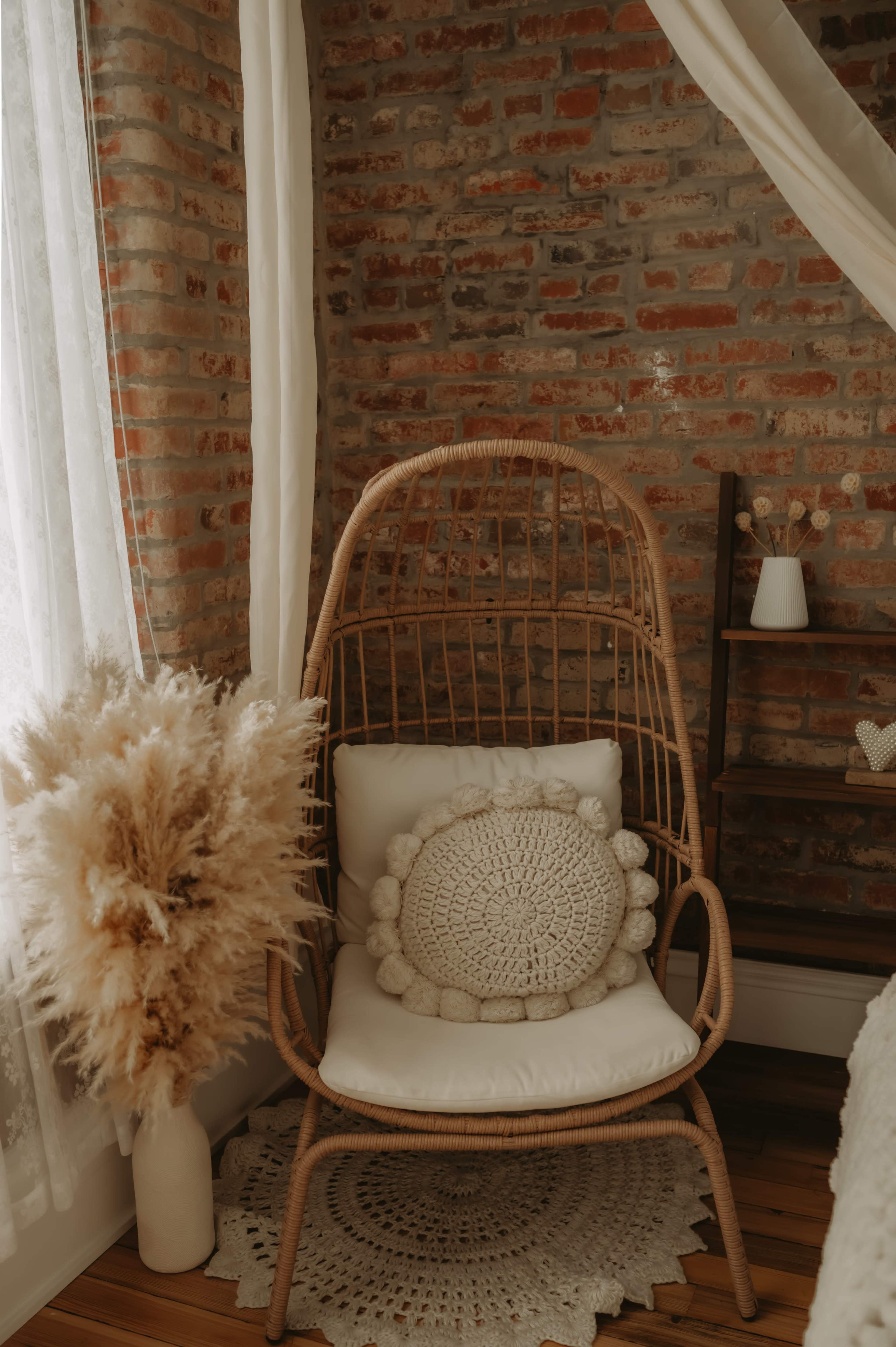 A wicker chair with a textured cushion is positioned beside a potted pampas grass arrangement against a brick wall.