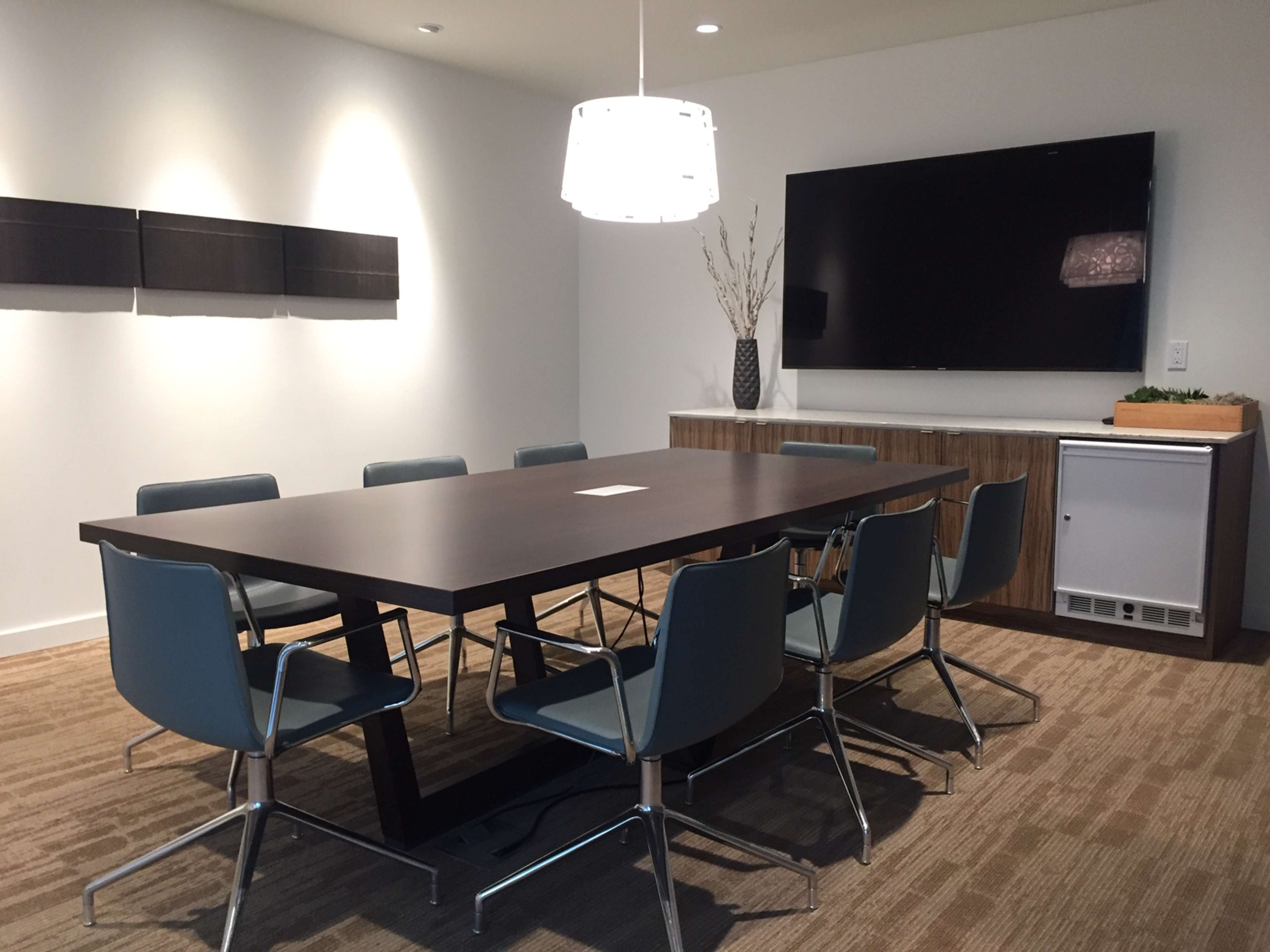 A conference room features a large rectangular table surrounded by six blue chairs, a wall-mounted television, and minimal decorative elements.