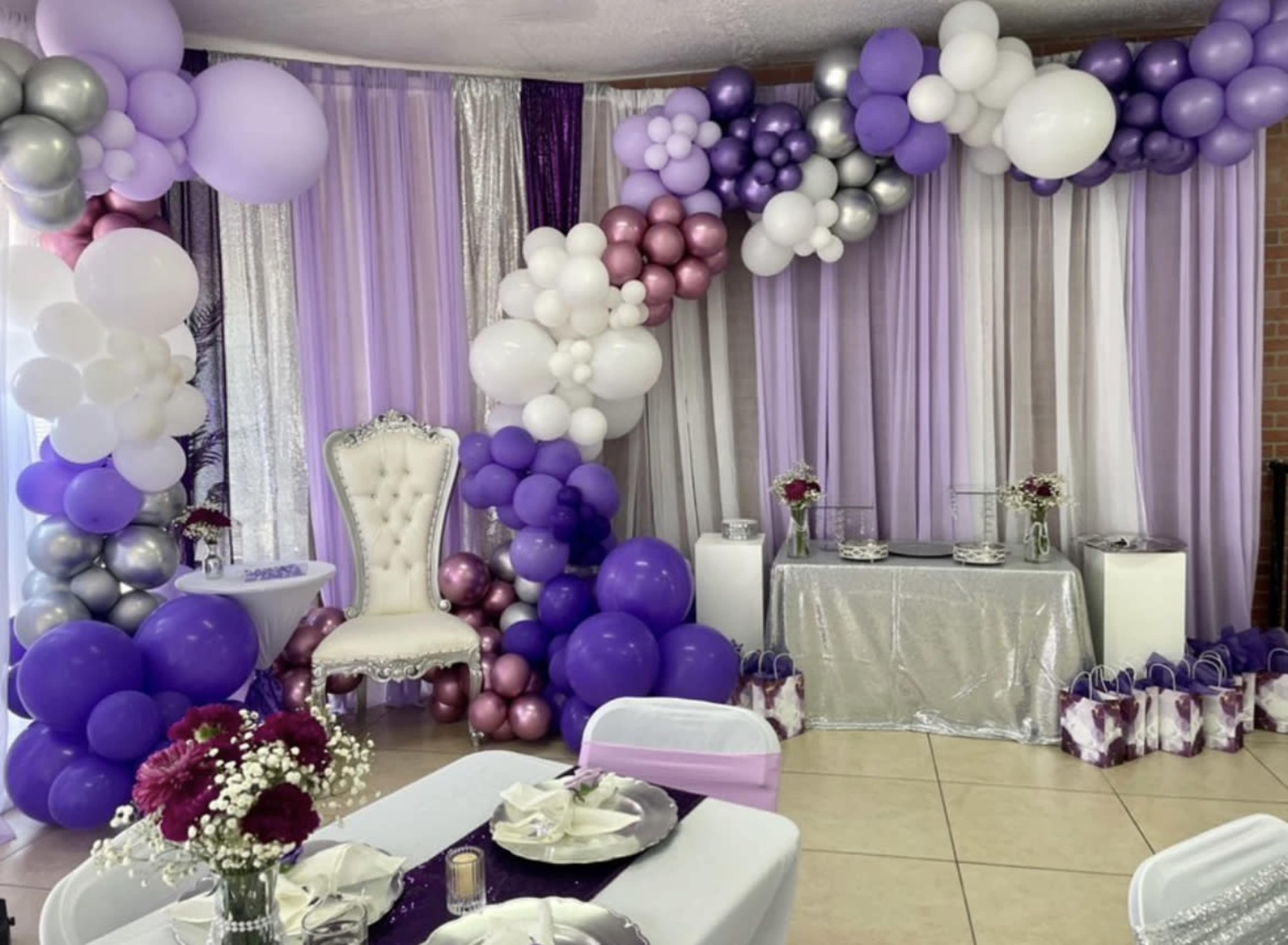 The image shows a decorated event space with a balloon arch in purple, silver, and white, a luxurious white throne chair, and a table set for a celebration.