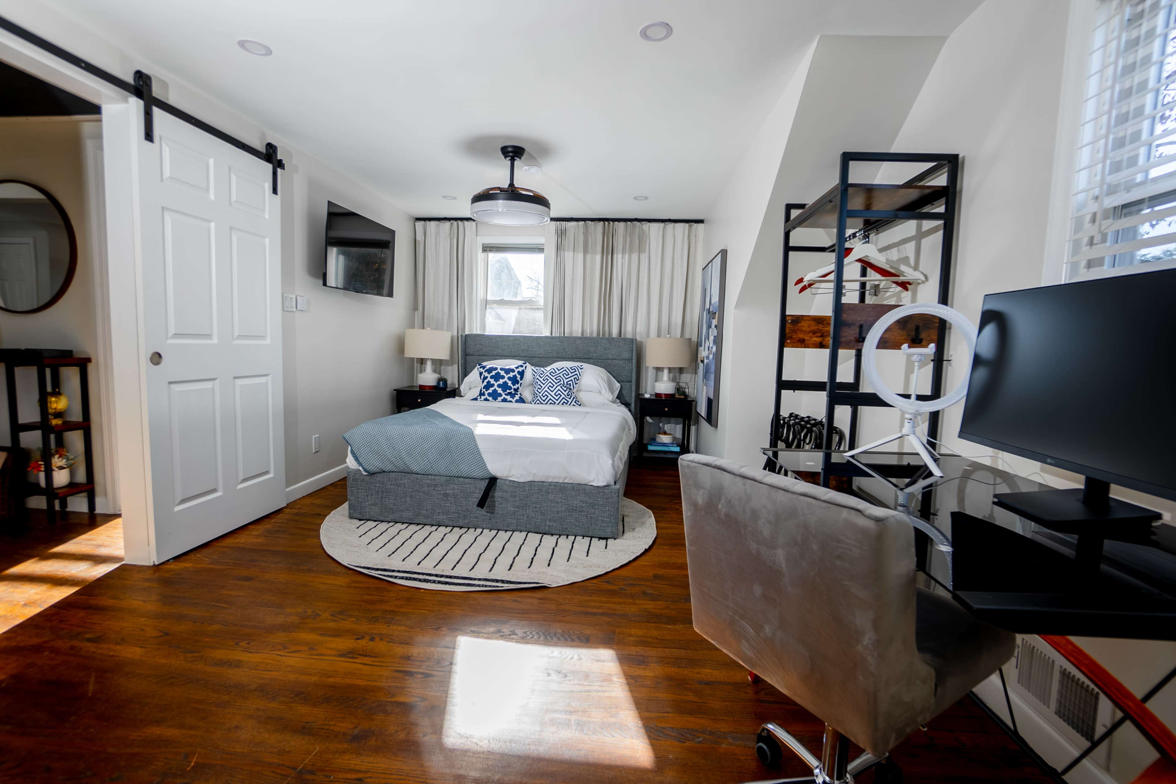 A well-arranged bedroom featuring a bed with white linens, a circular rug, a desk with a chair, and a window with sheer curtains.