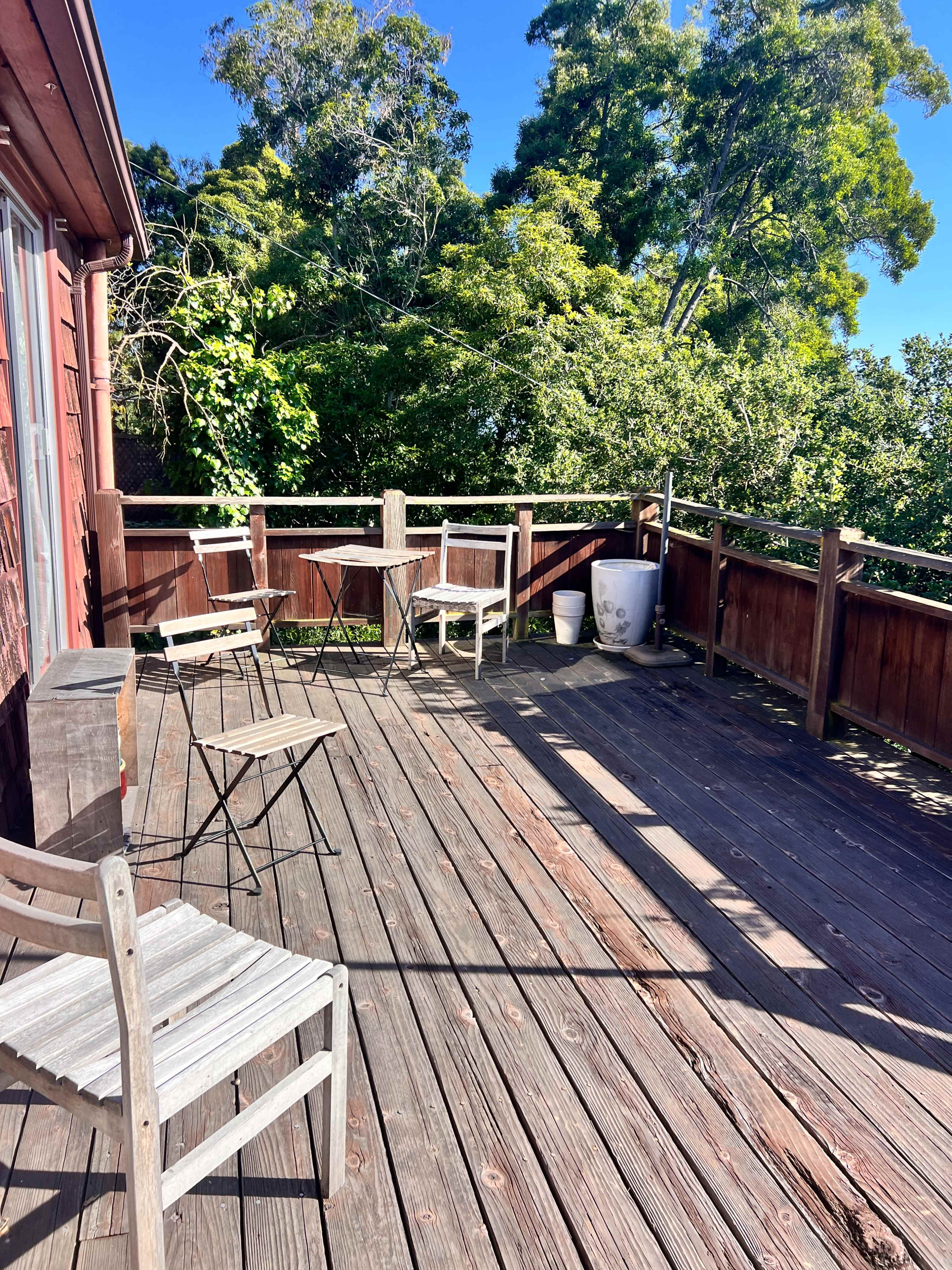 The image shows a wooden deck with several metal chairs and a table, surrounded by lush green trees.