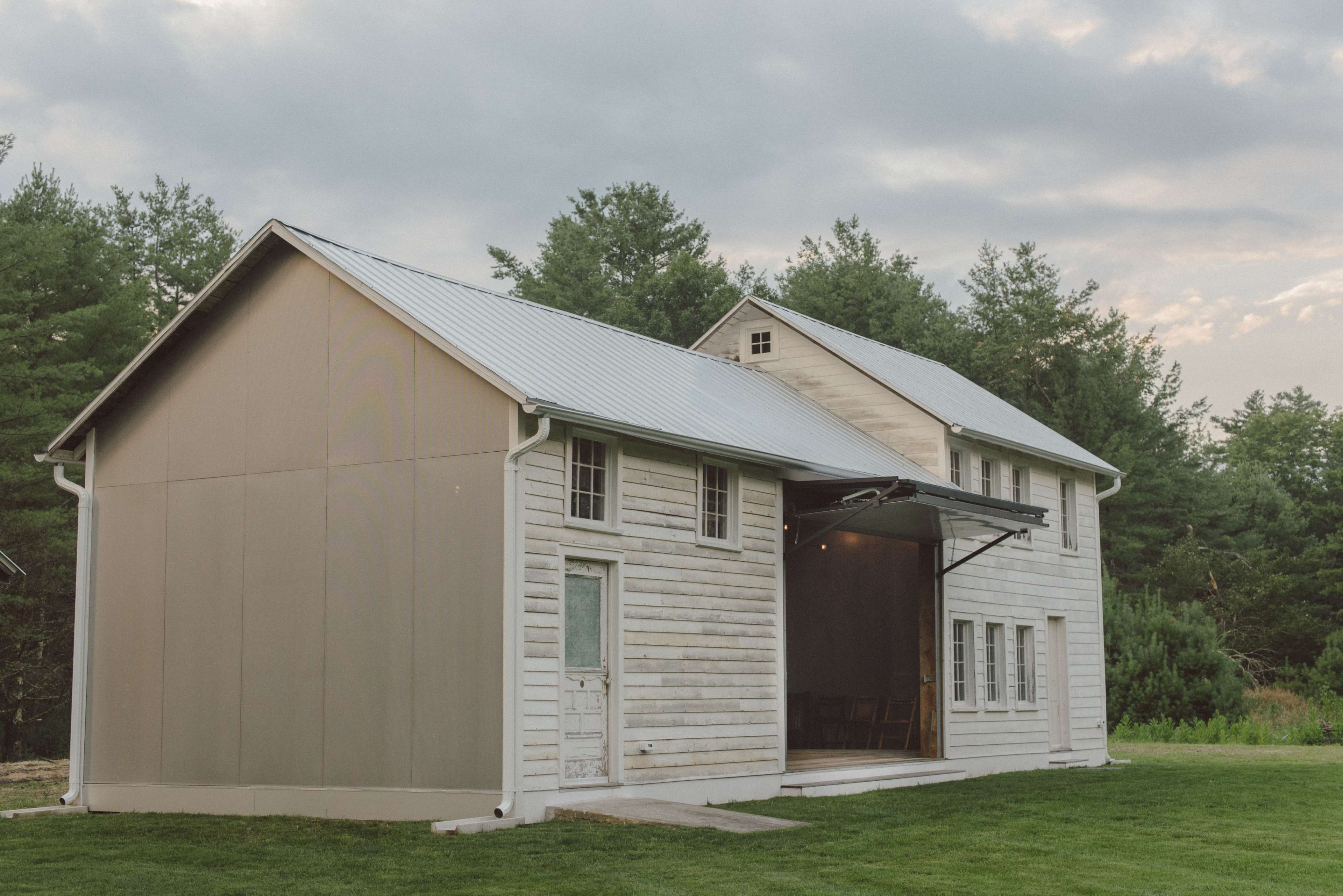 A two-story, modern farmhouse with a metal roof, situated on a grassy area surrounded by trees.