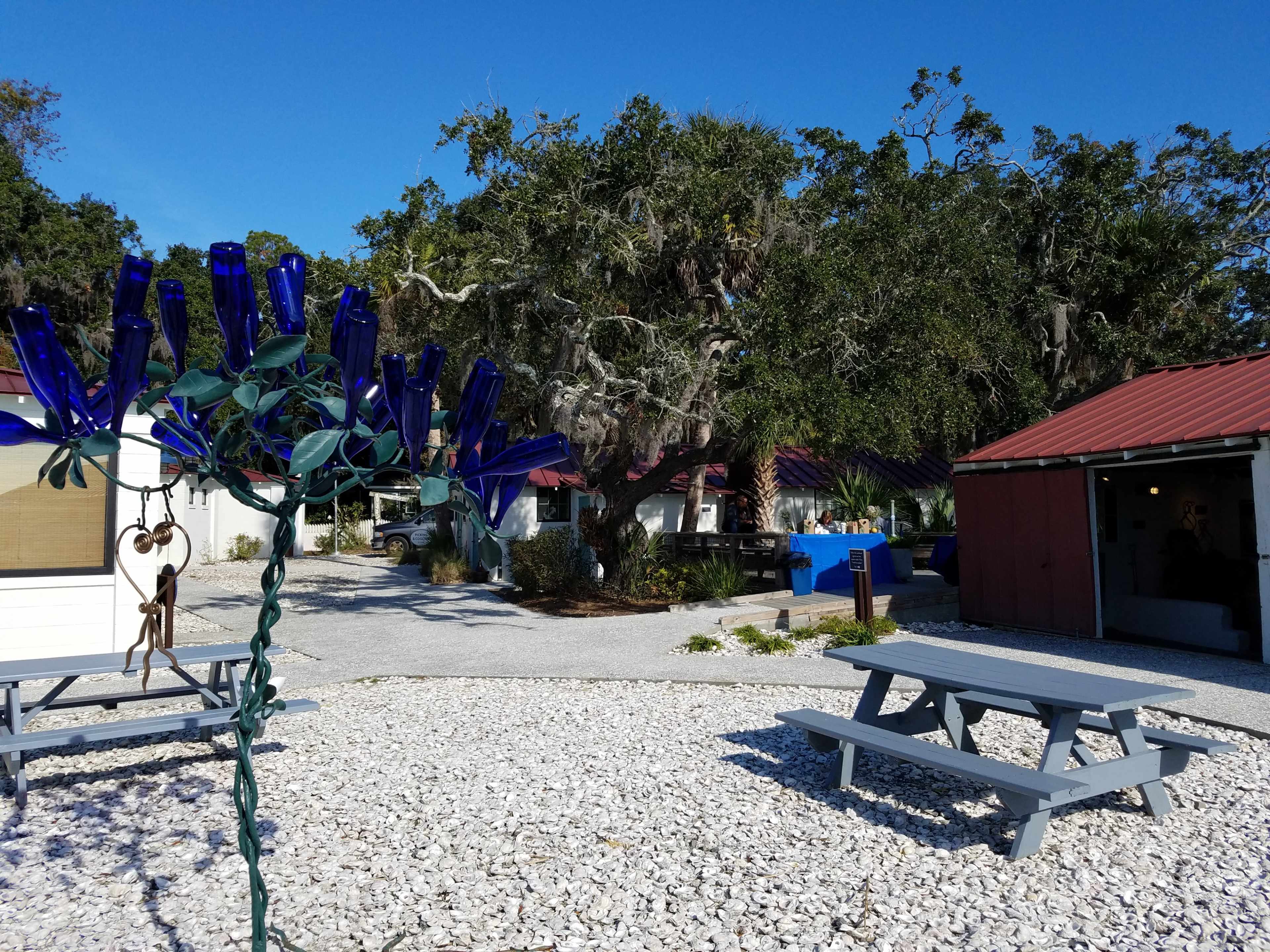 A courtyard features picnic tables and a tree decorated with blue glass bottles, surrounded by buildings with red roofs and large trees.