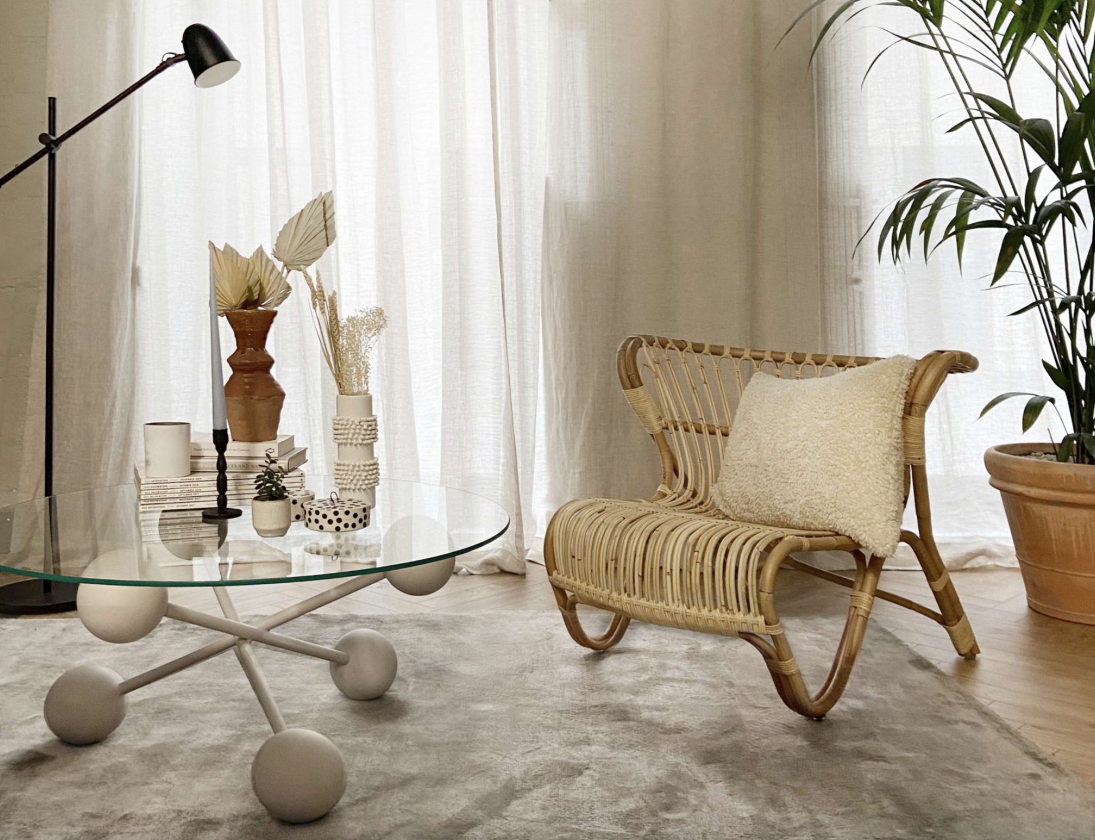 A rattan chair and a glass coffee table with decorative items sit on a rug in a well-lit room with sheer curtains and a potted plant.