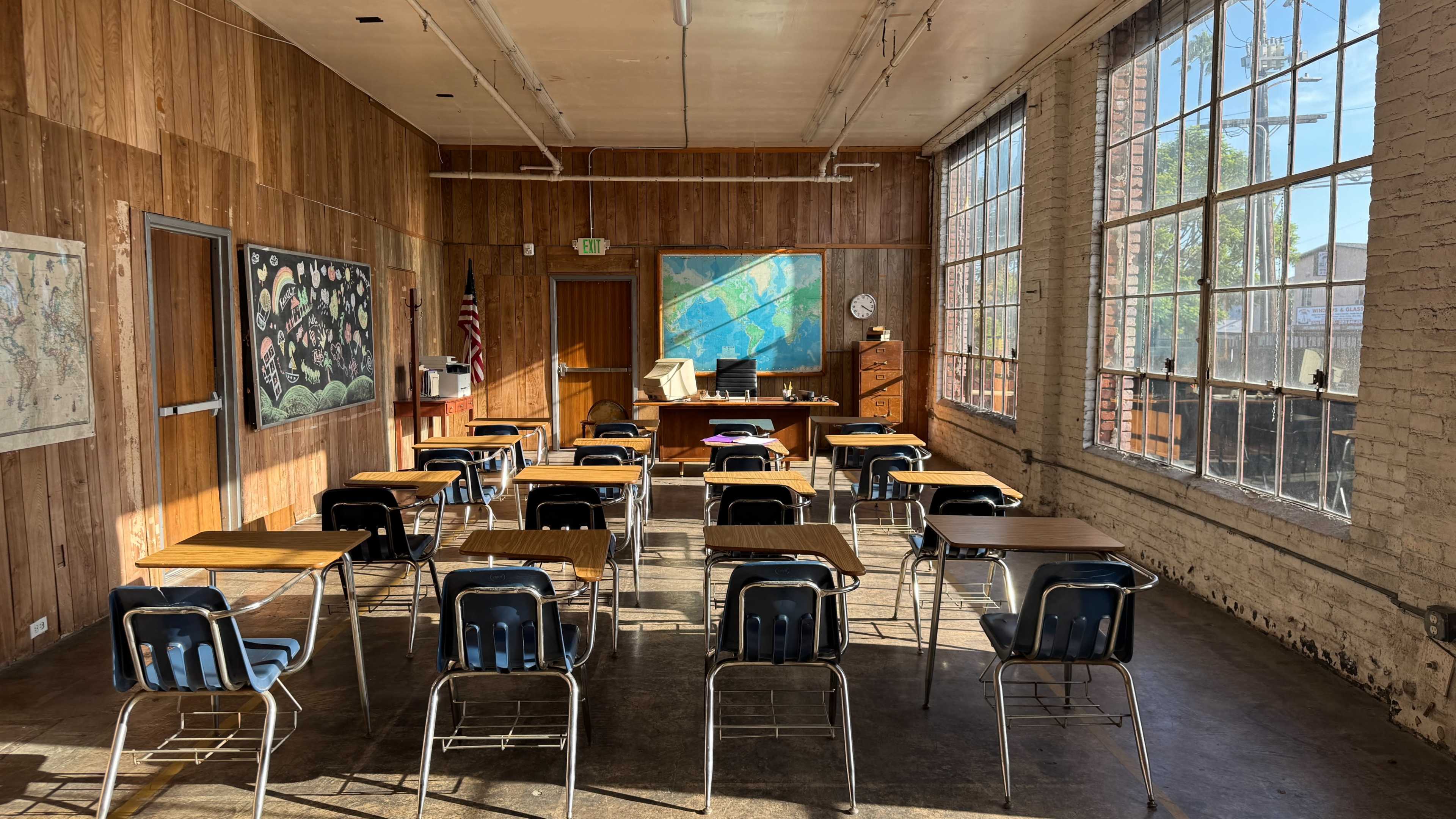 The image shows a classroom with wooden walls, a map on the wall, and rows of desks facing a teacher's desk near large windows.