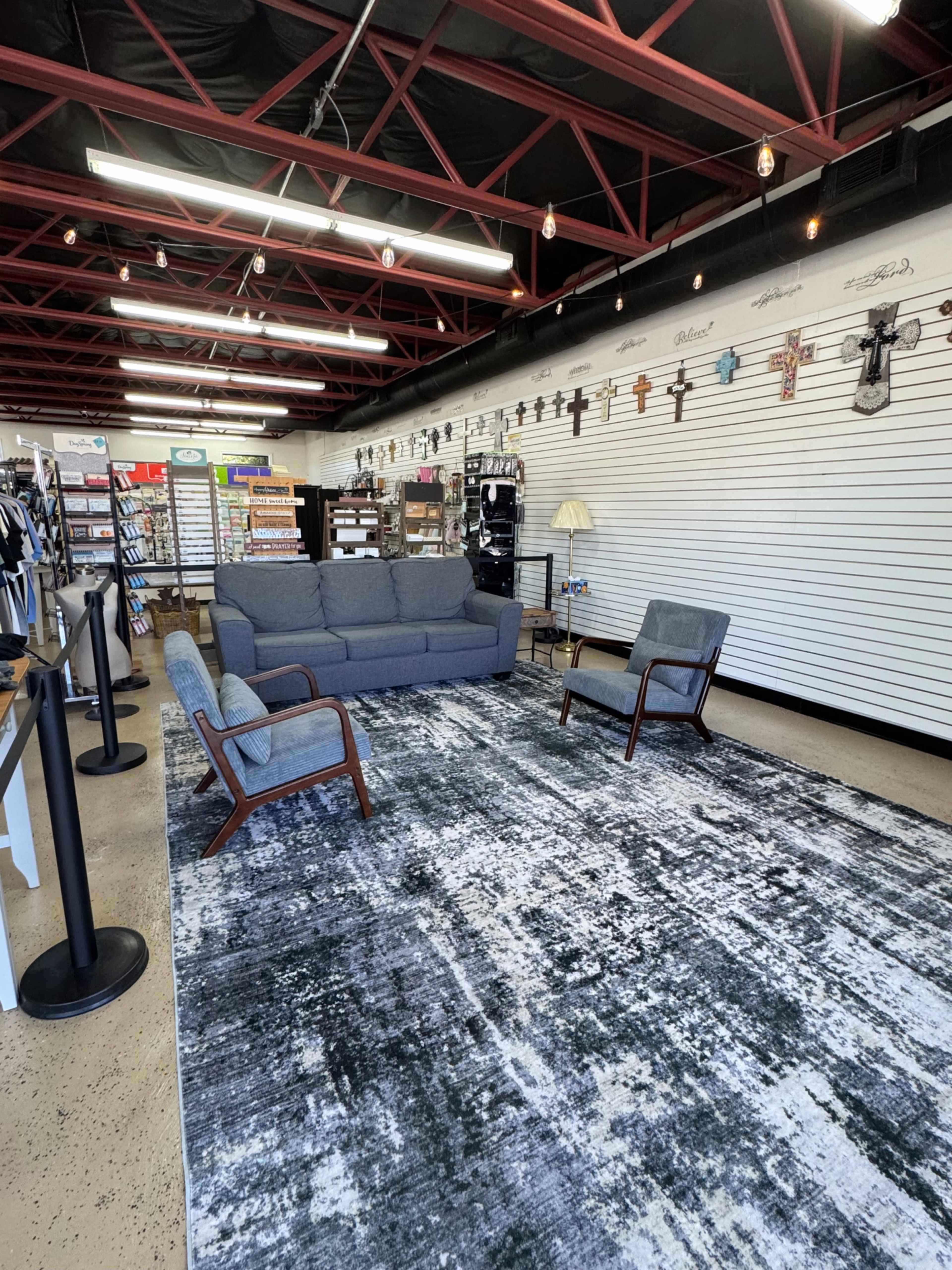 The image shows a spacious retail area featuring a gray sofa and two chairs set on a patterned rug, with merchandise displayed on white walls behind them.