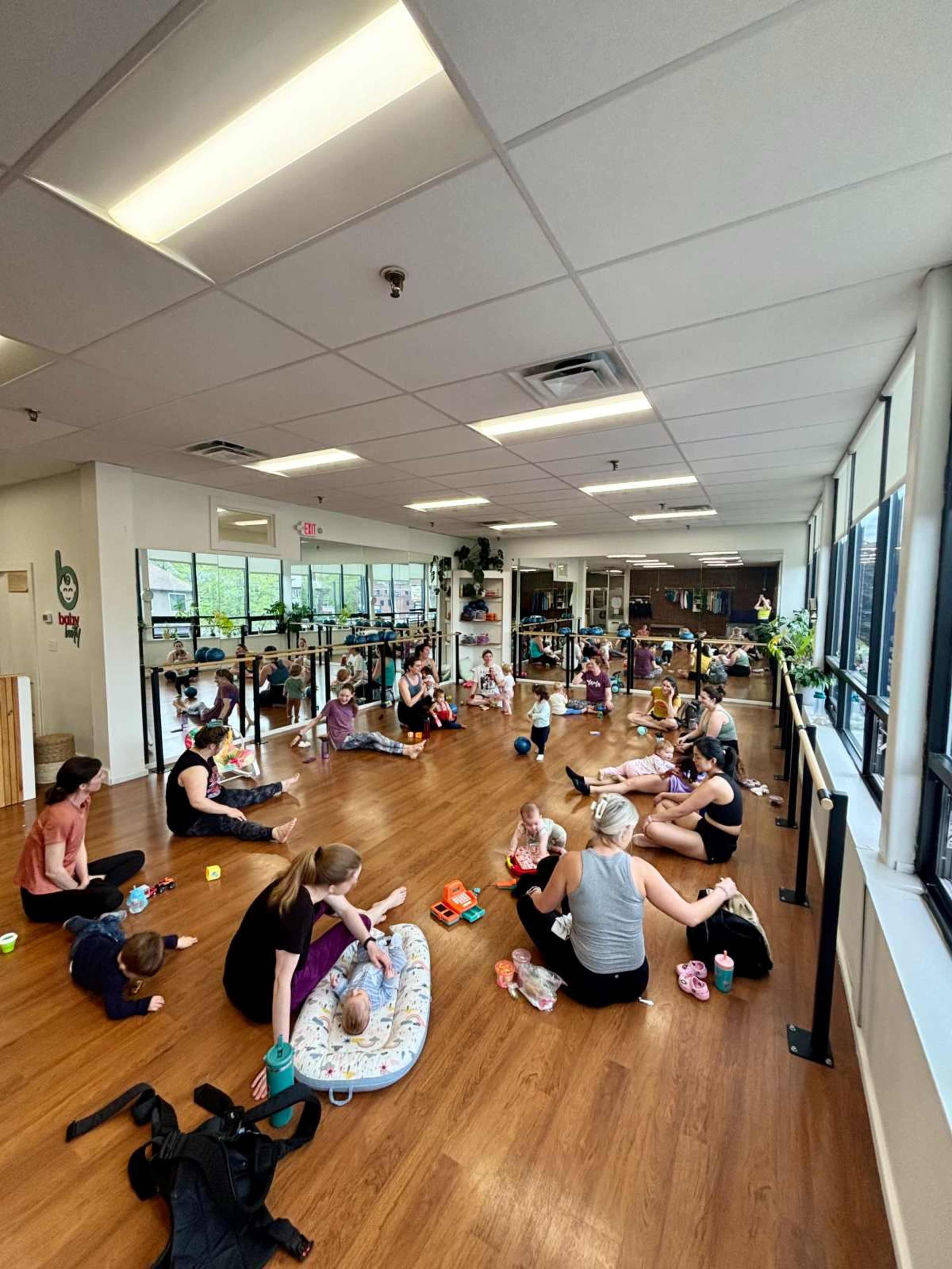A group of parents and their young children participate in a class in a well-lit studio with wooden flooring and large windows.