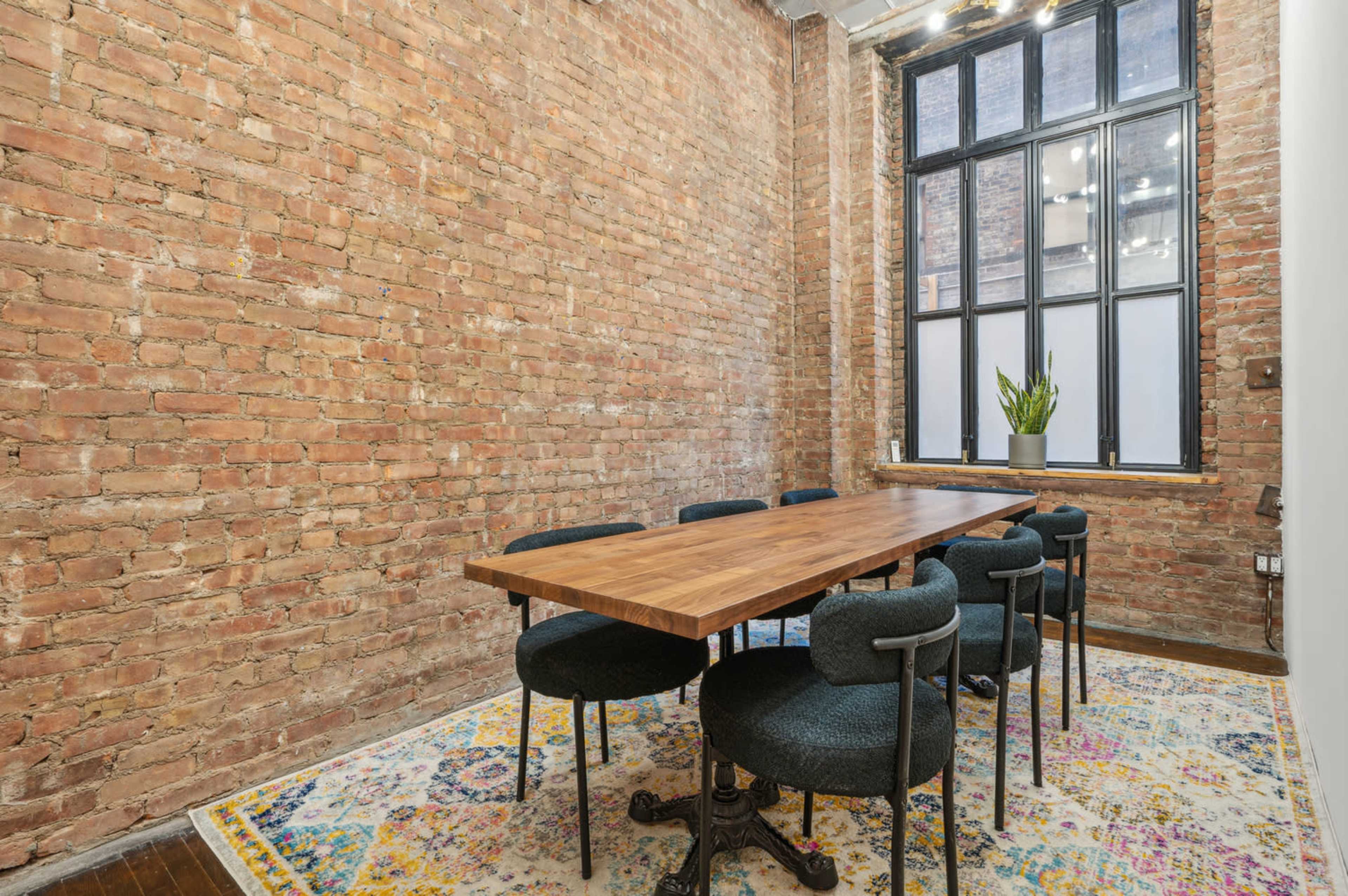 A rustic meeting room features a large wooden table surrounded by six chairs on a colorful area rug, with exposed brick walls and a large window.