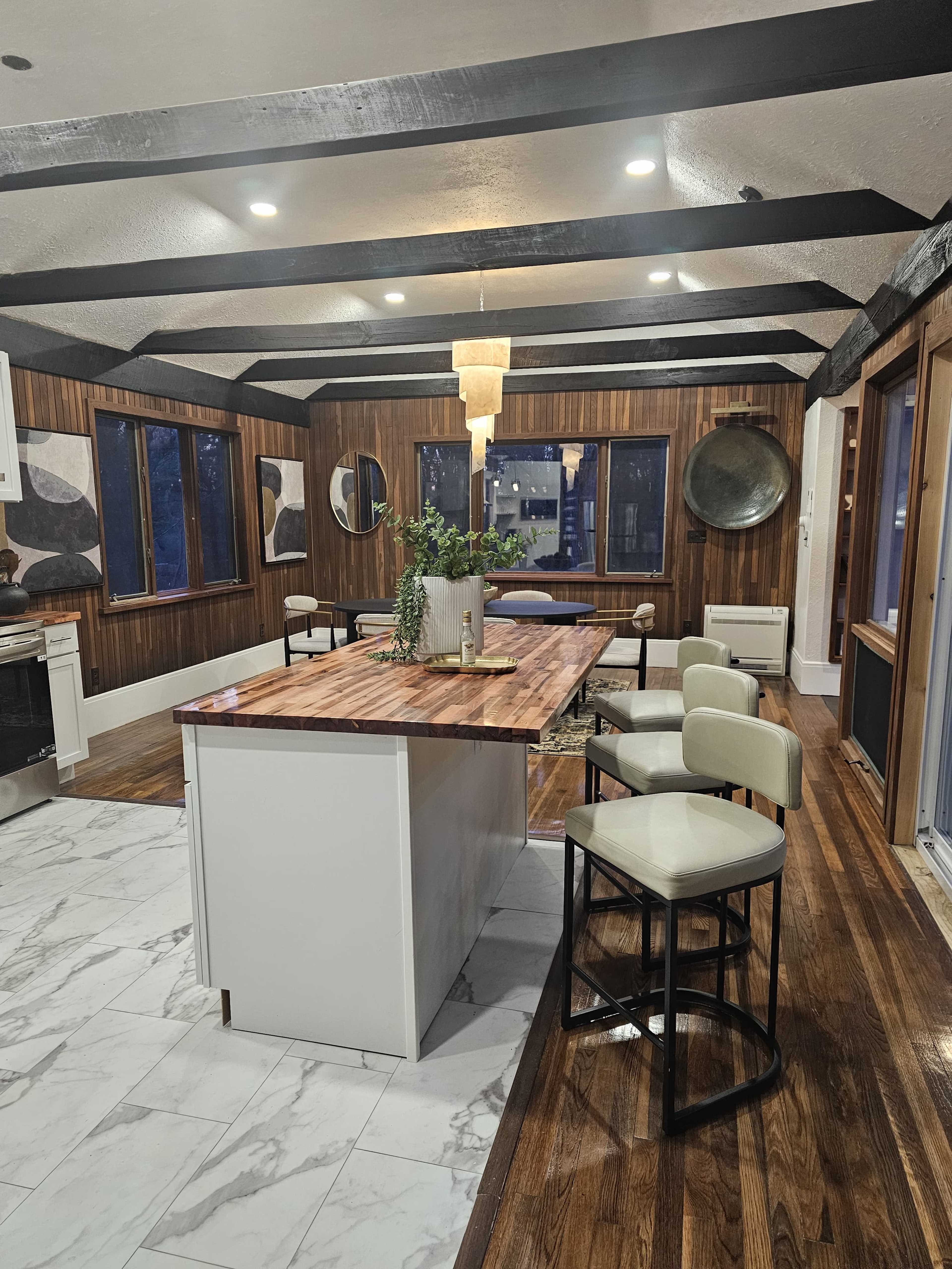 The image shows a modern kitchen featuring wood-paneled walls, a large central island with a wooden countertop, and a combination of marble and wooden flooring.