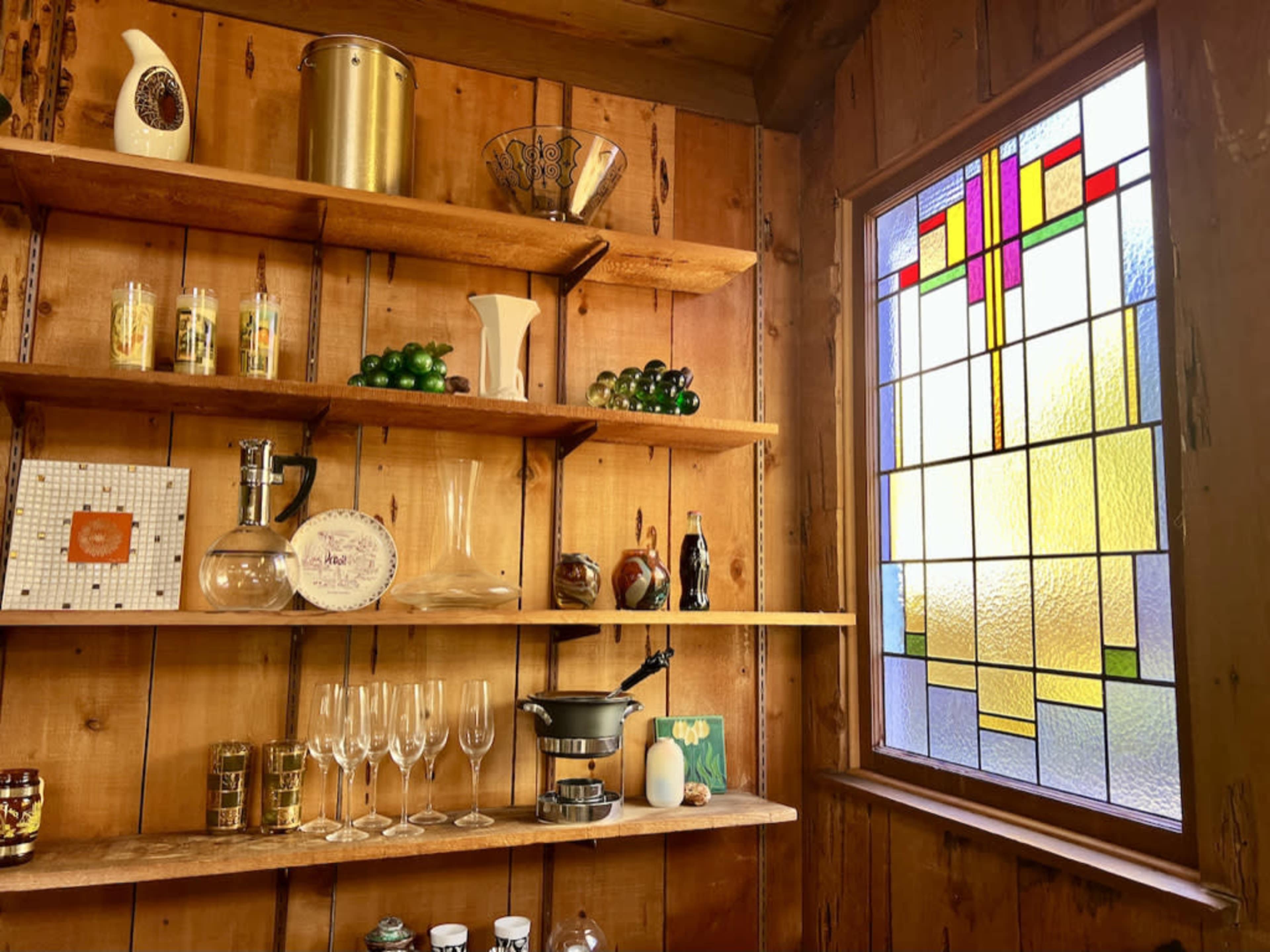 A wooden wall with shelves holding various glassware and decorative items, adjacent to a stained glass window.