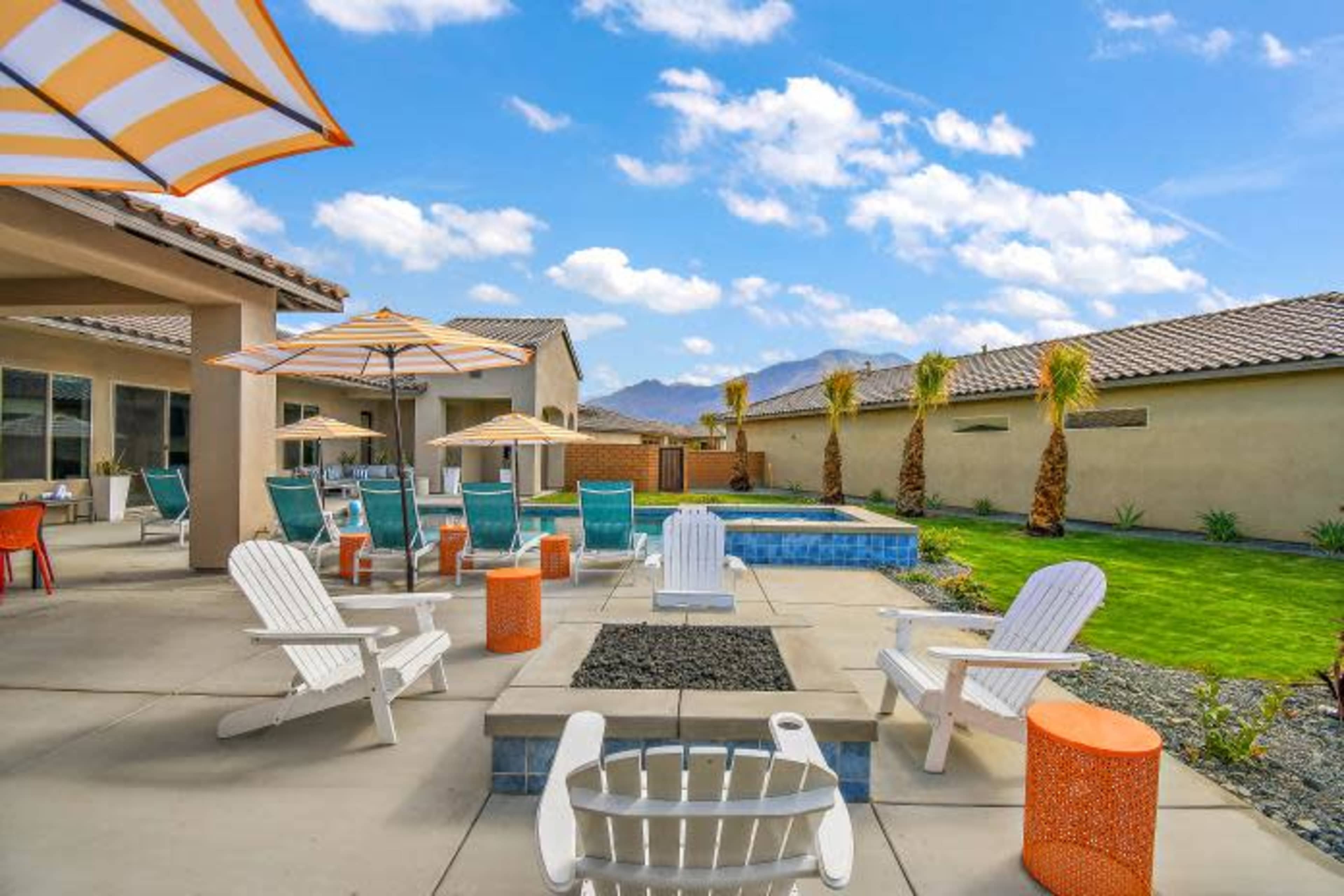 A spacious outdoor patio area with lounge chairs, umbrellas, a small pool, and palm trees, set against a backdrop of blue skies and distant mountains.