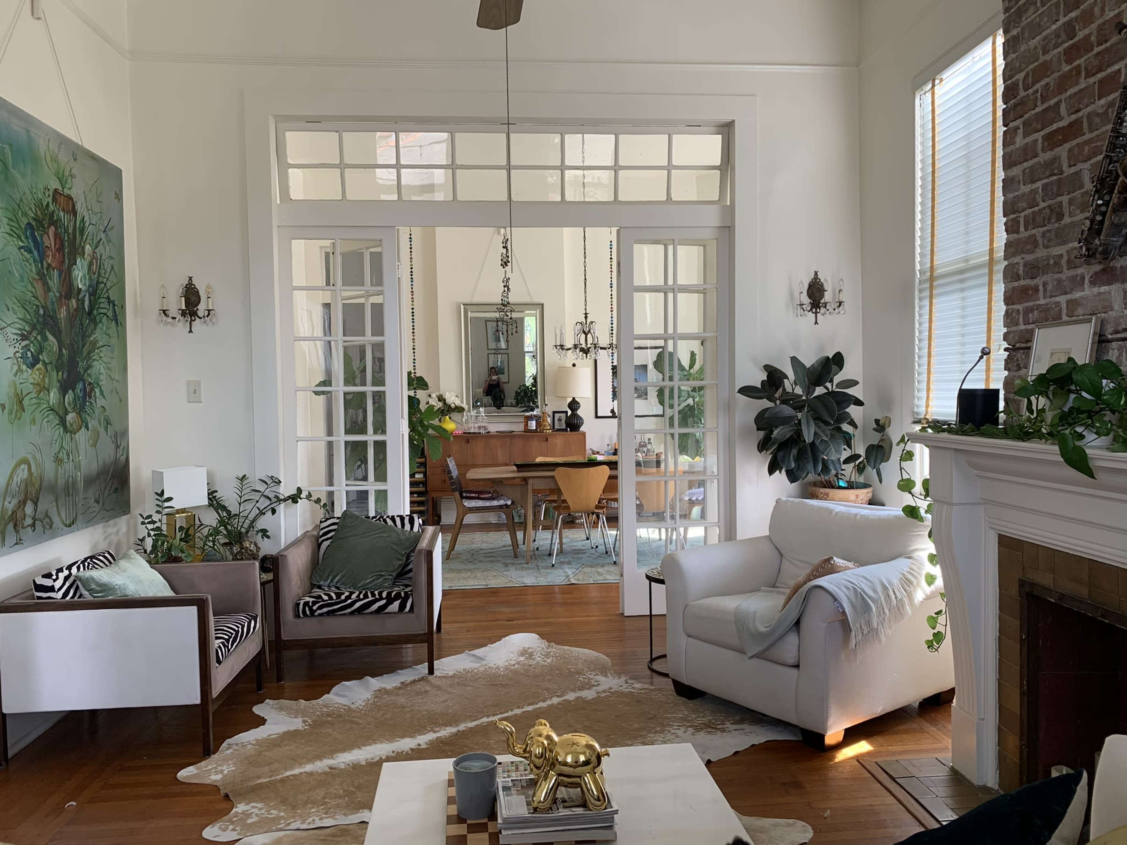 A bright living room with large windows, a mix of plants, and a view into a dining area featuring a wooden table and chairs.