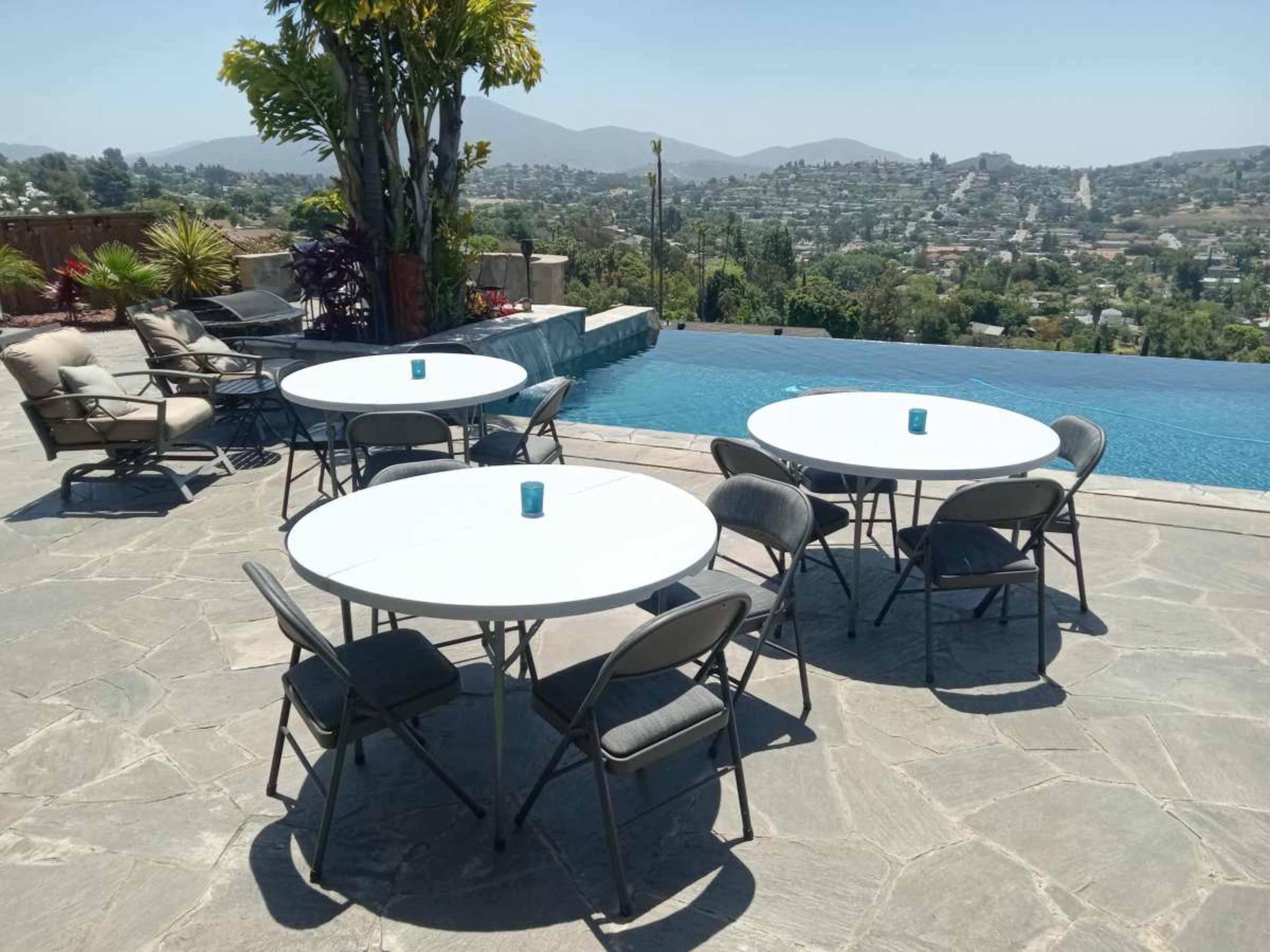 A patio area features several circular tables surrounded by chairs, overlooking an infinity pool and a distant hillside.