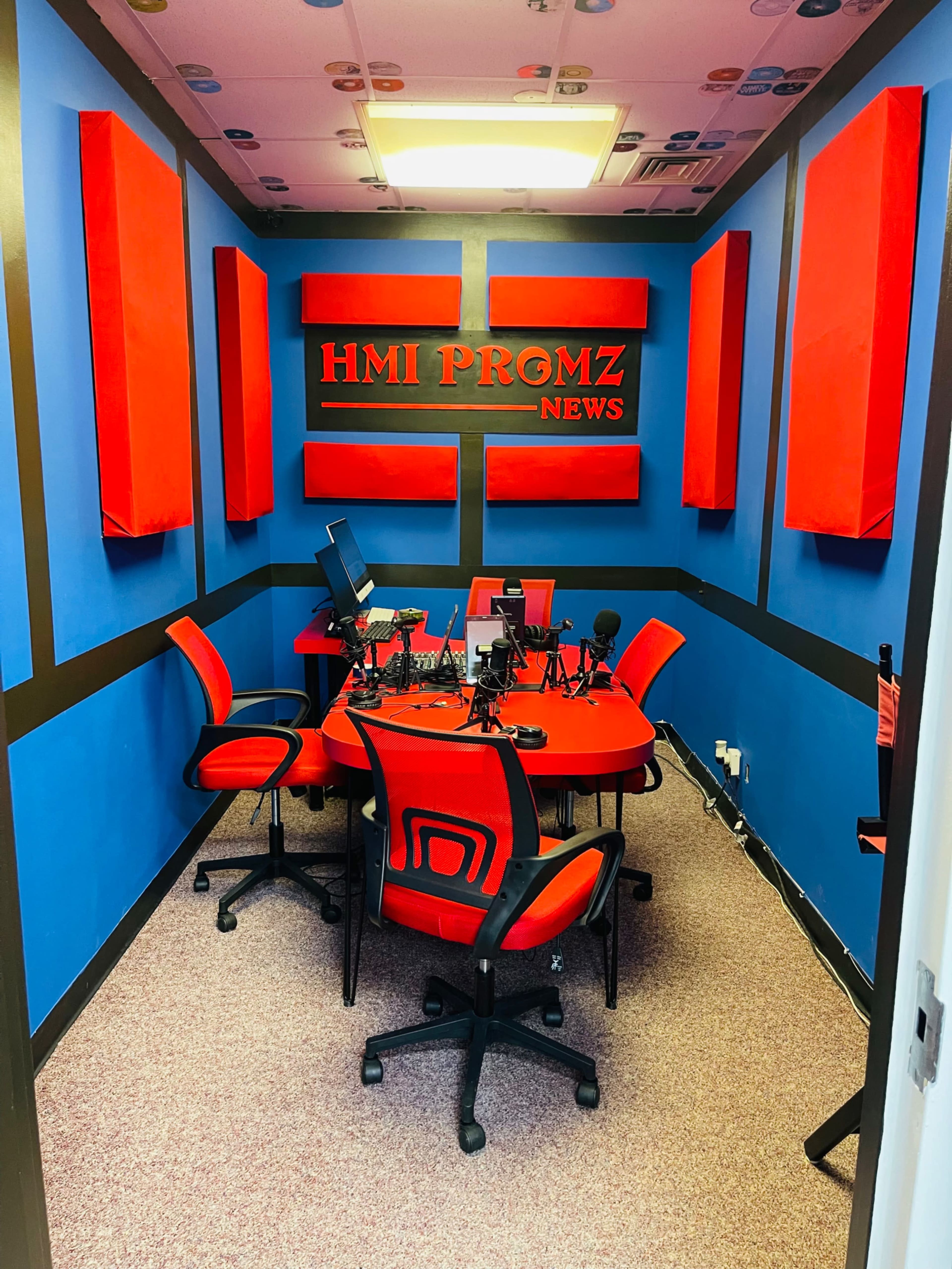 A small news studio featuring a circular red table, black microphones, and red chairs, surrounded by blue walls and acoustic panels.