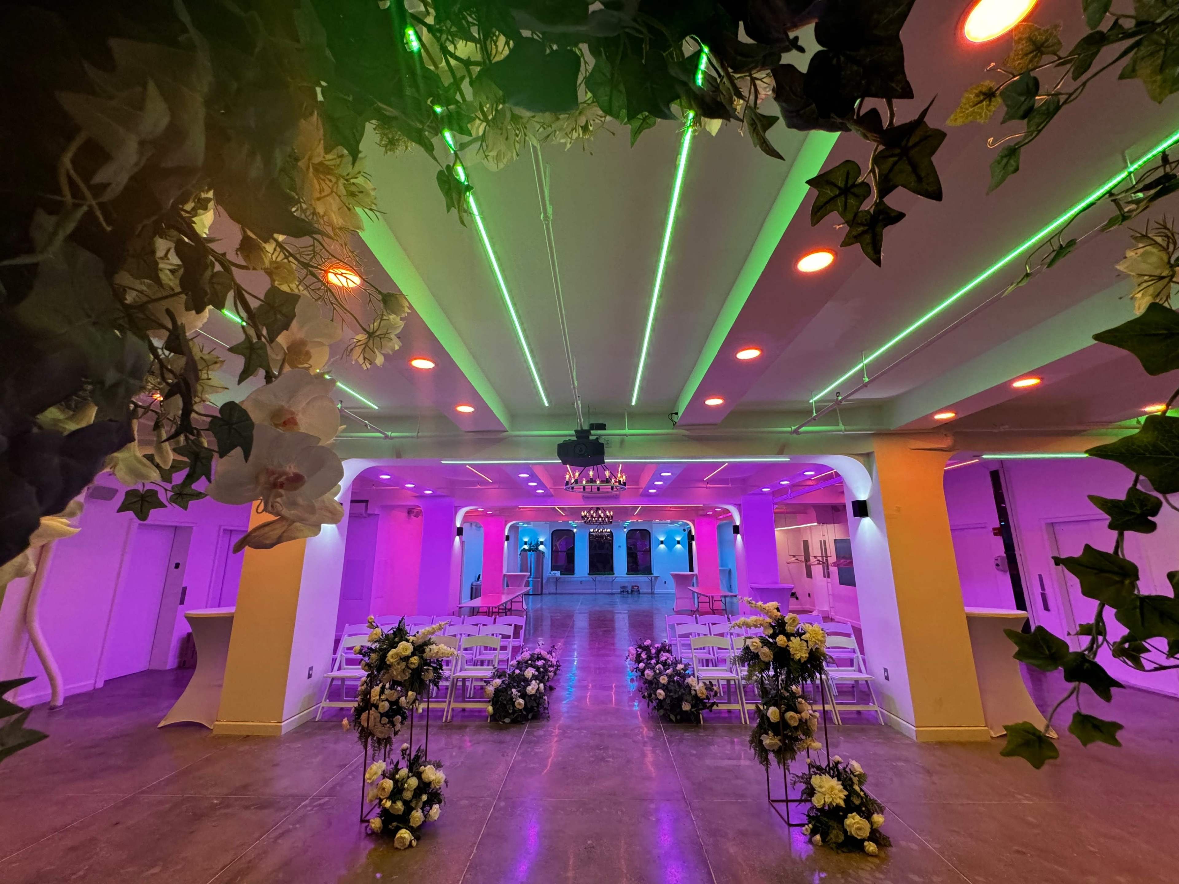 A decorated indoor space features rows of white chairs and floral arrangements, illuminated by colorful lights.