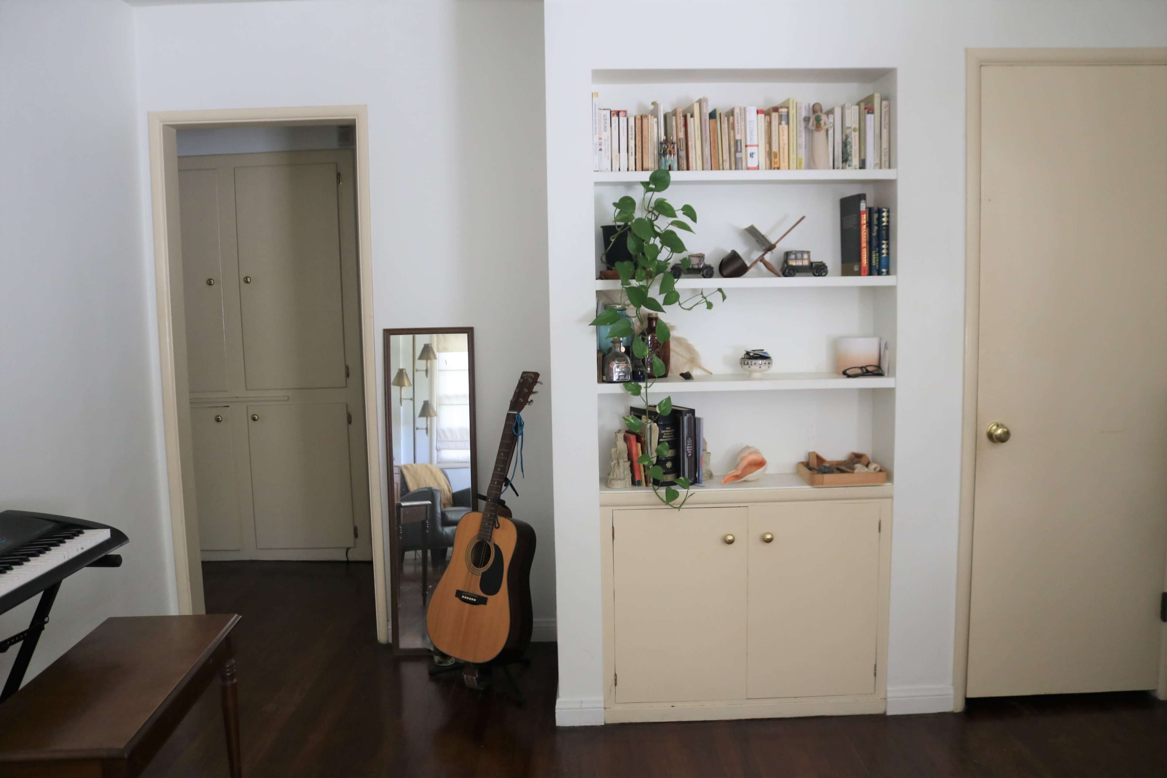 A room with a wooden floor, a bookshelf with books and decorative items, an acoustic guitar leaning against a cabinet, a mirror, and a doorway leading to another room.